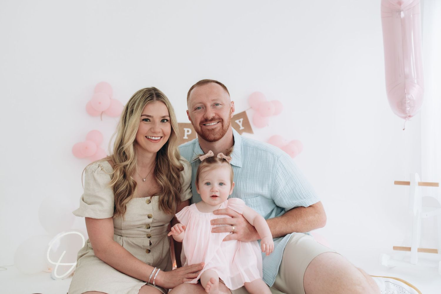 A family of three in soft neutral clothing sits together and smiles on a white bed.