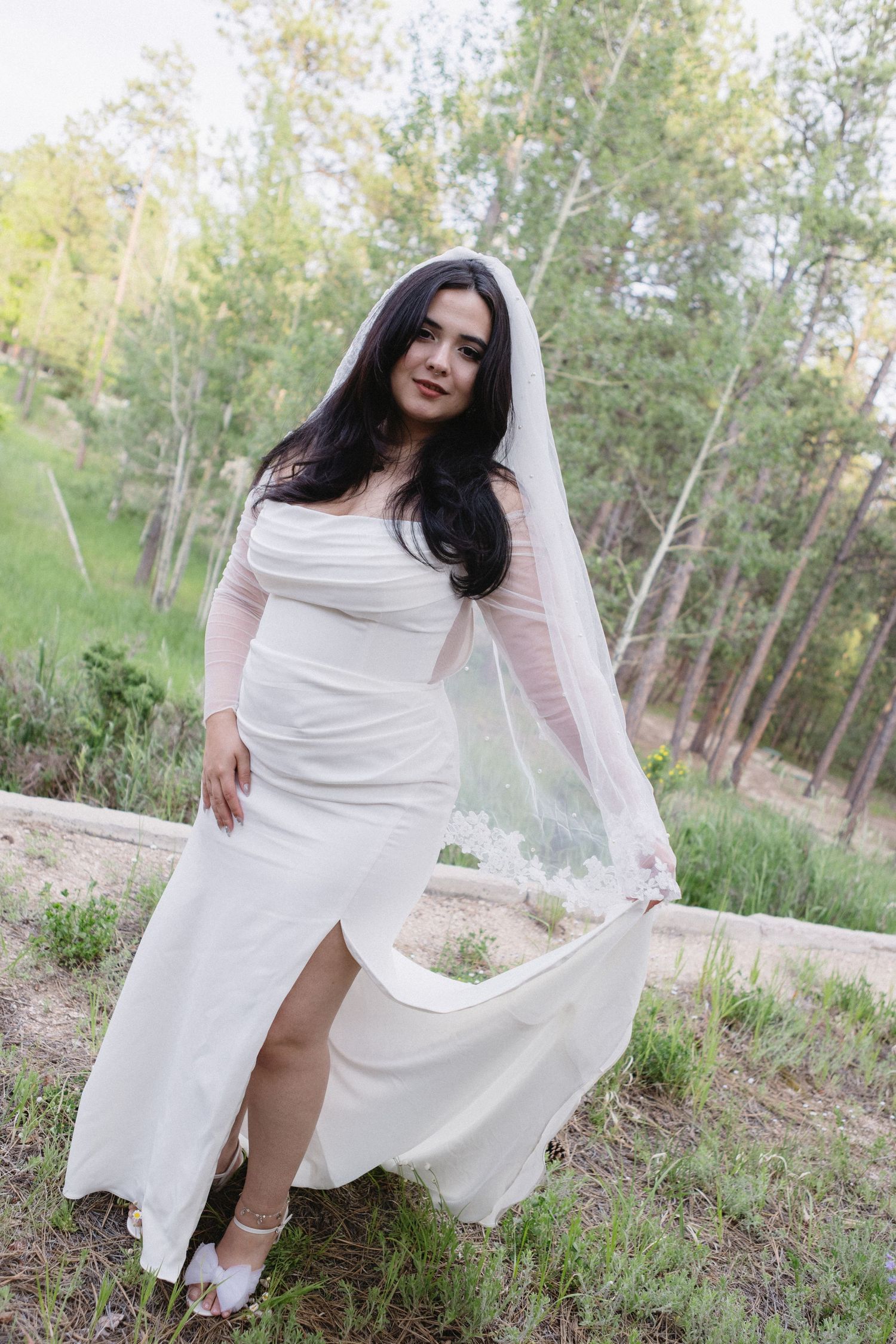 A bride in a white wedding dress posing in a wooded outdoor setting with tall green trees in the background.