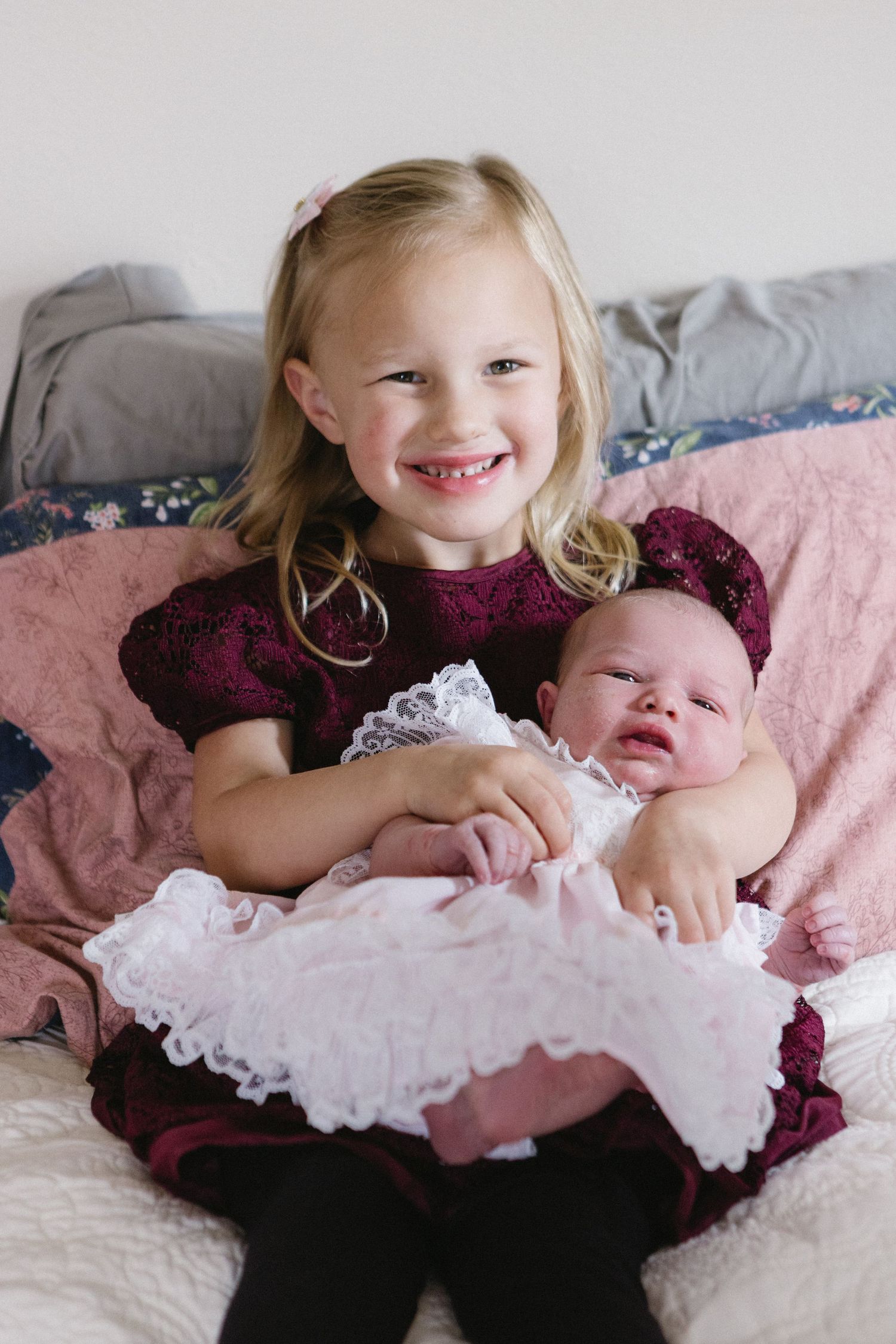 Two children in coordinating burgundy outfits sit on pink bedding, with a sweet embrace between siblings.