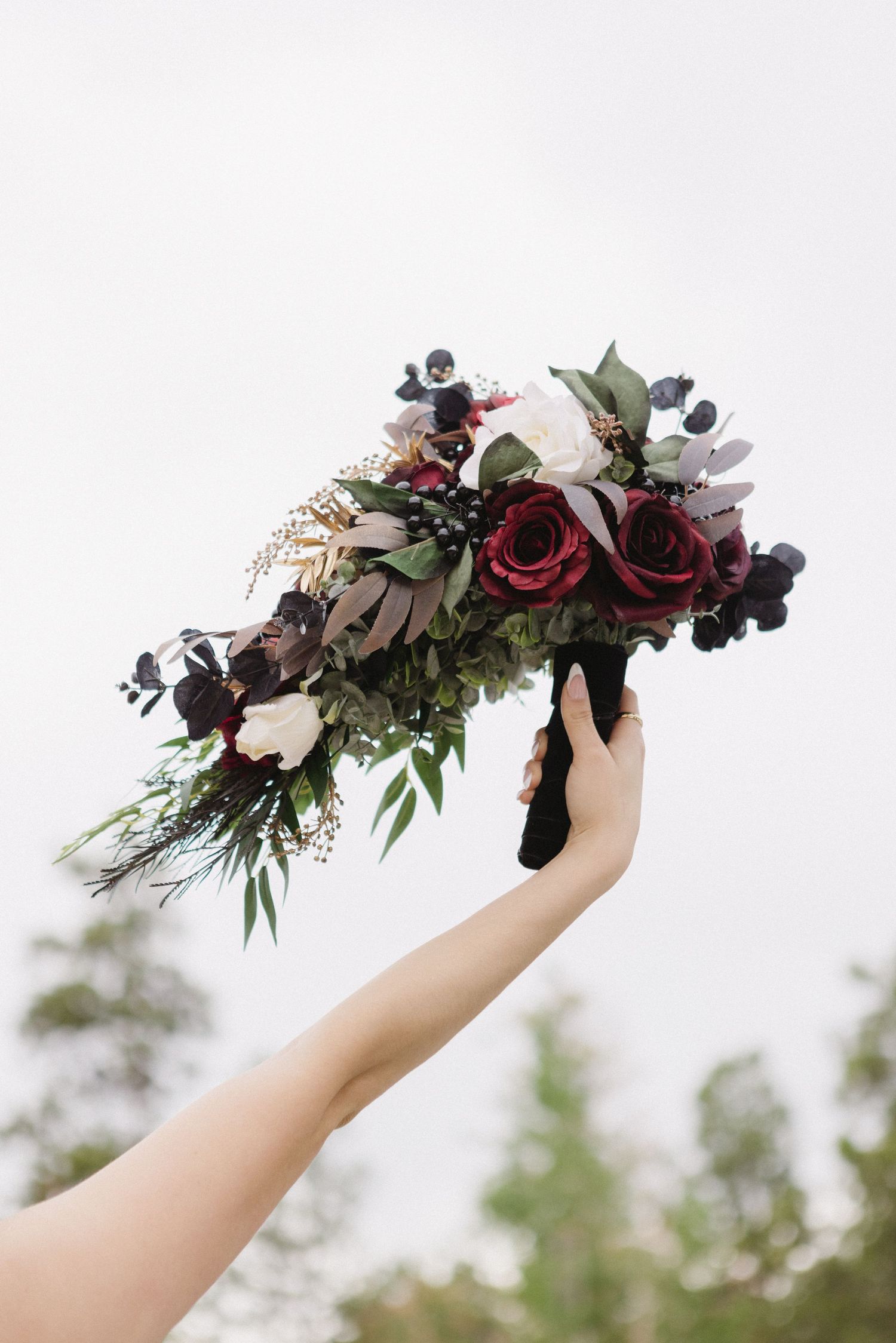 A cascading floral bouquet with burgundy roses and dark berries held up against a bright sky background.