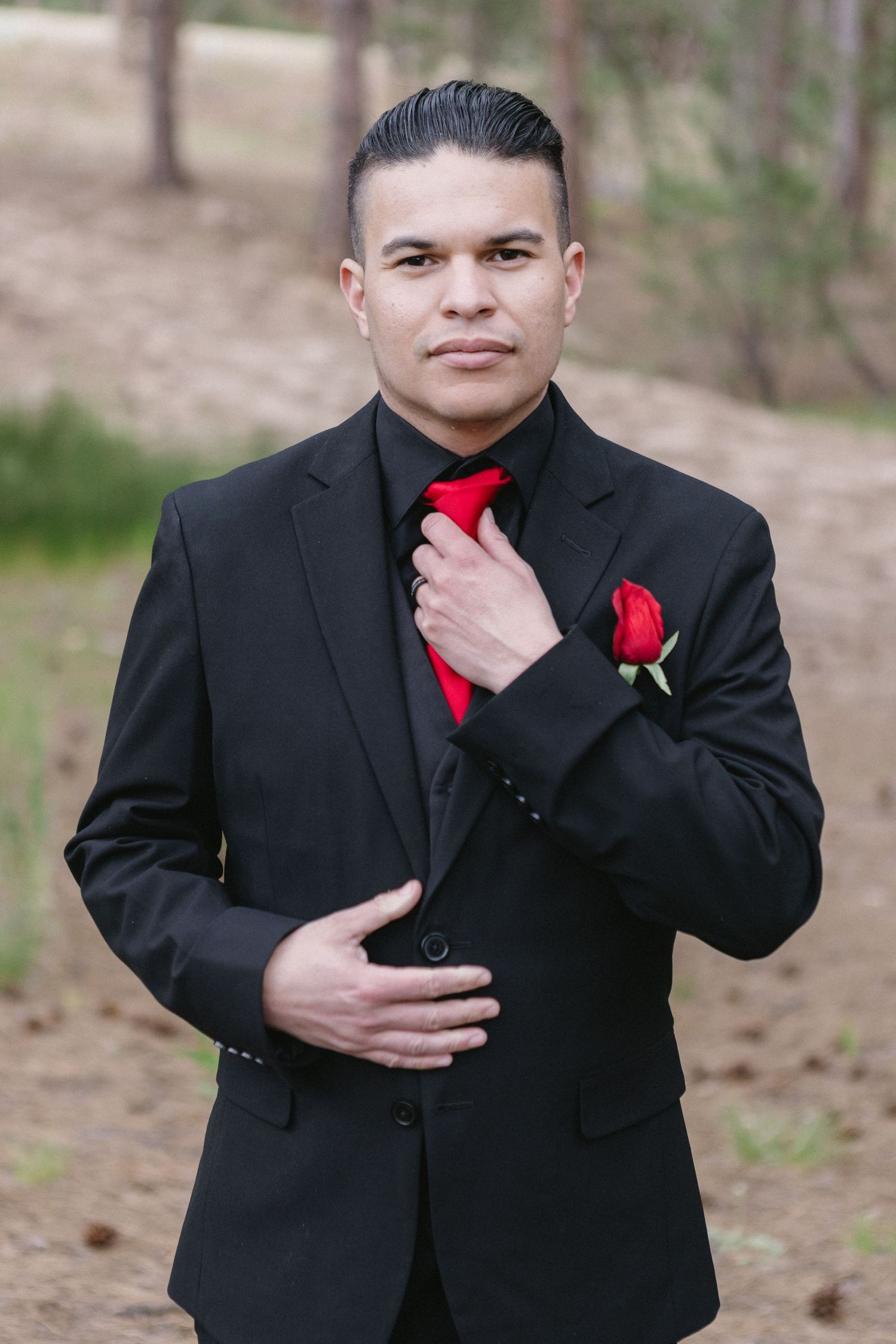 Formal portrait of someone wearing a black suit with red tie and boutonniere adjusting their tie outside in a natural setting.
