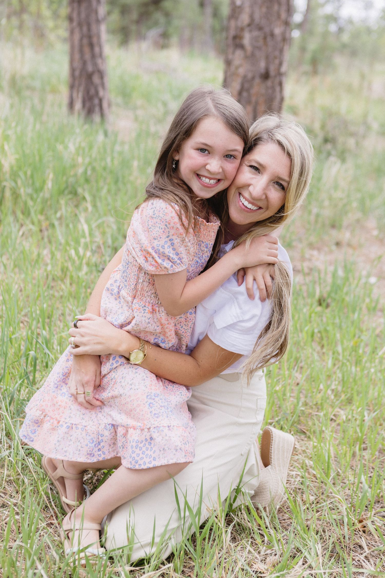 A touching outdoor photoshoot shows two people hugging and sitting together in tall grass on a sunny day.
