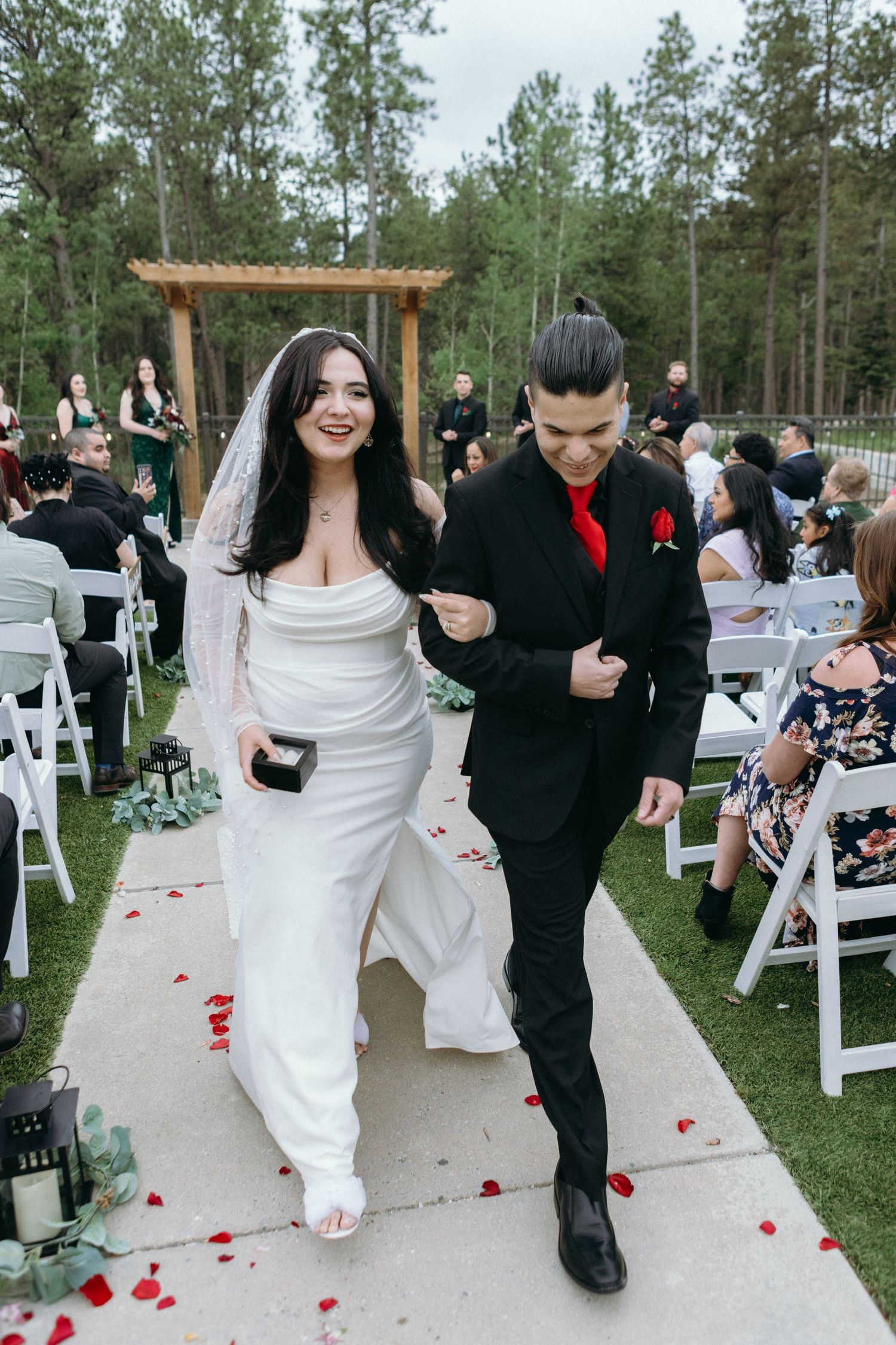 Newlyweds walk down outdoor aisle surrounded by white chairs and pine trees while guests celebrate with rose petals.