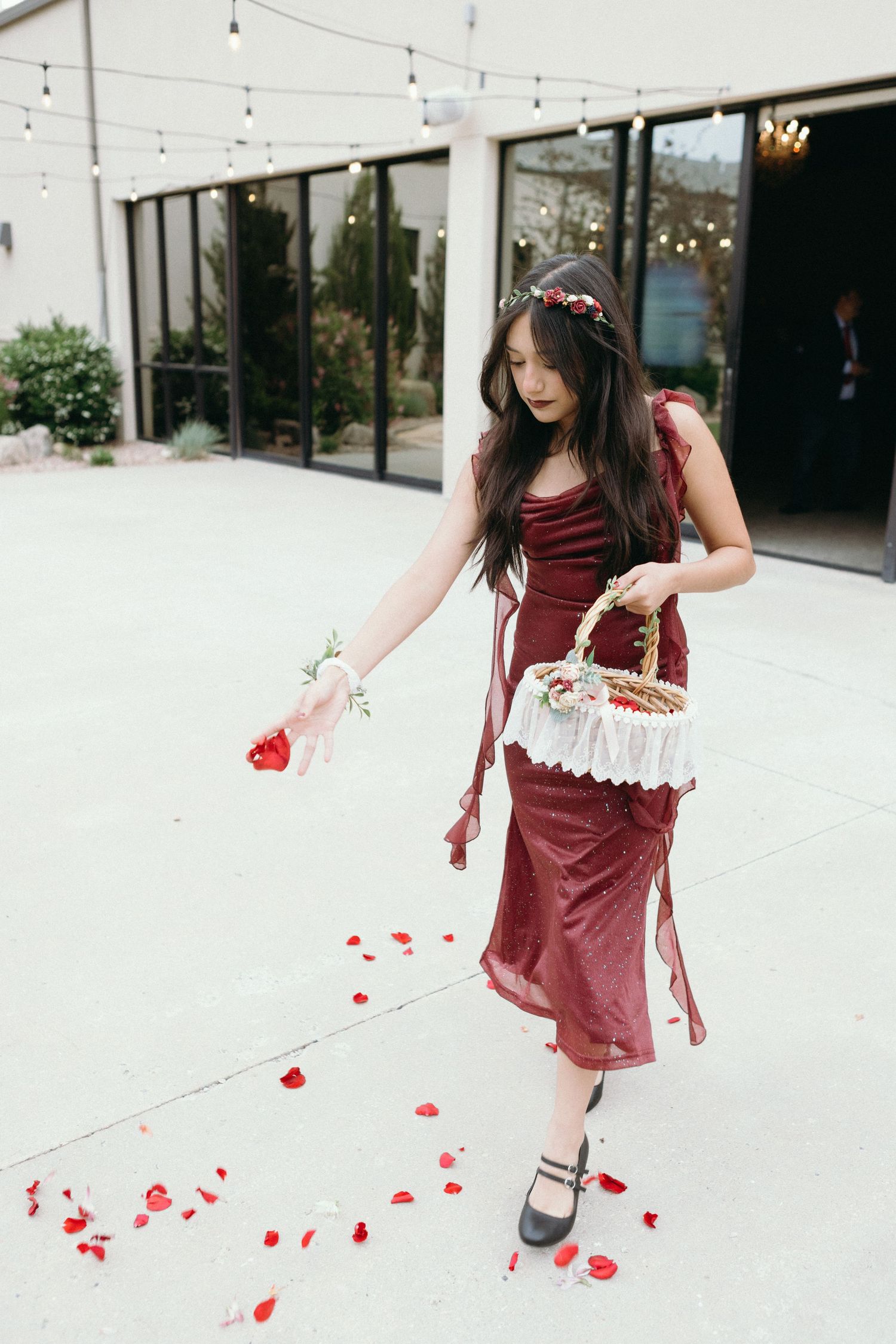 Person in burgundy velvet dress scattering rose petals on white patio with twinkling lights above.