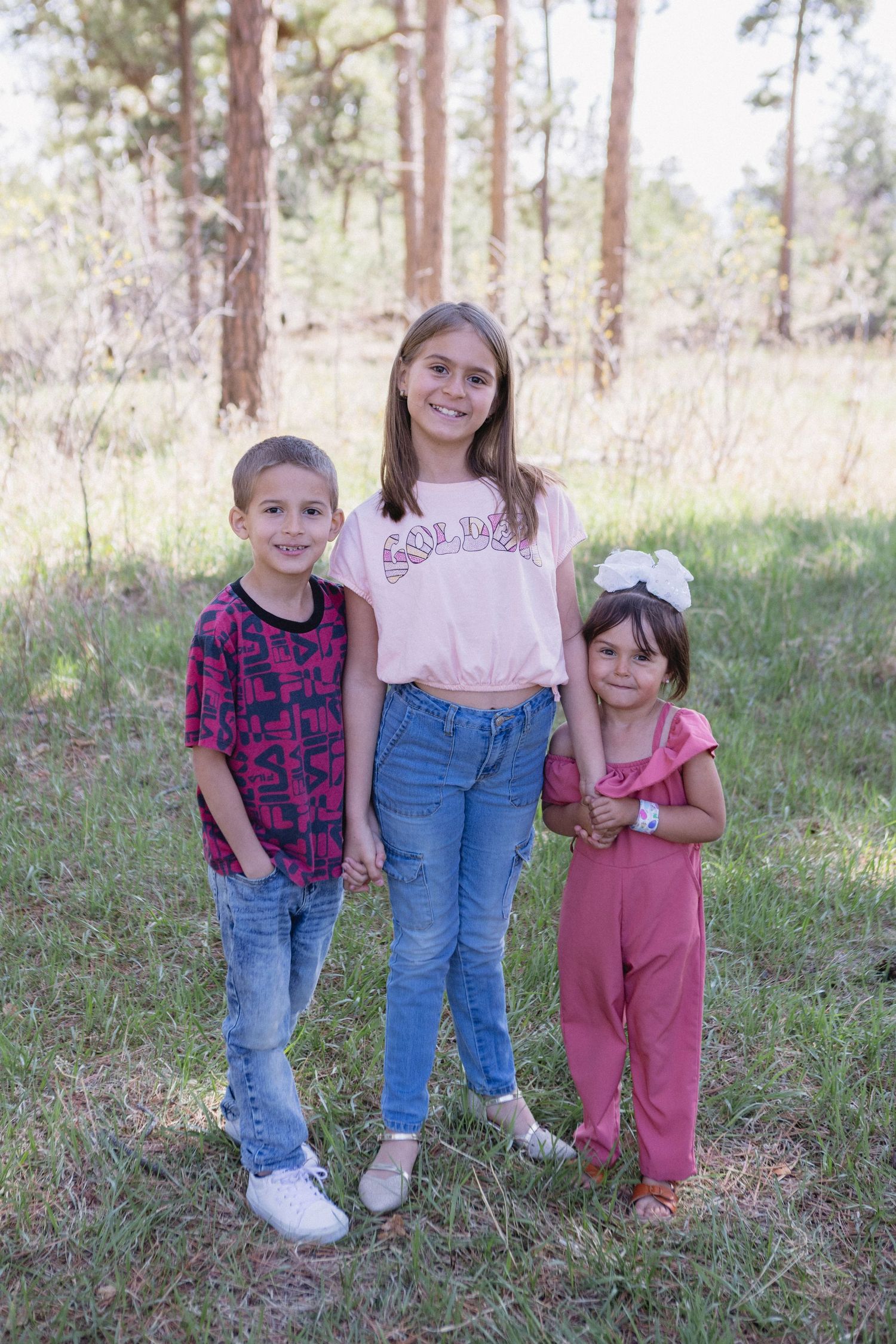 Three siblings standing together in a wooded outdoor setting wearing casual clothing on a sunny day.