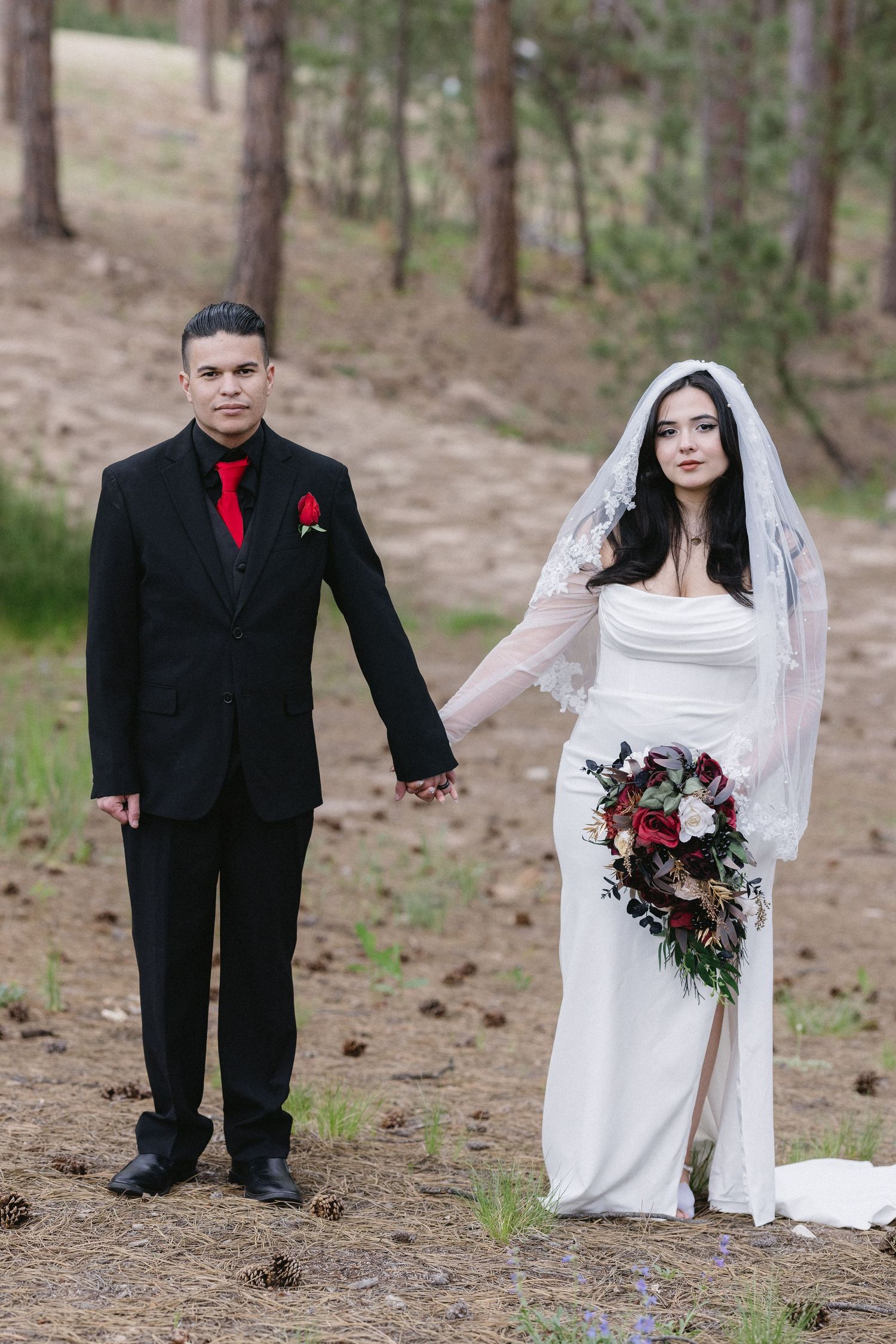 A wedding couple in black and white attire poses on a forest path with a red and burgundy floral bouquet.