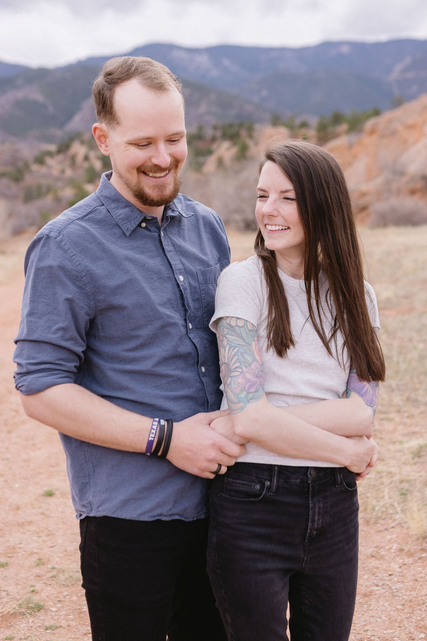 A smiling couple embraces on a mountain trail with scenic Colorado foothills in the background.