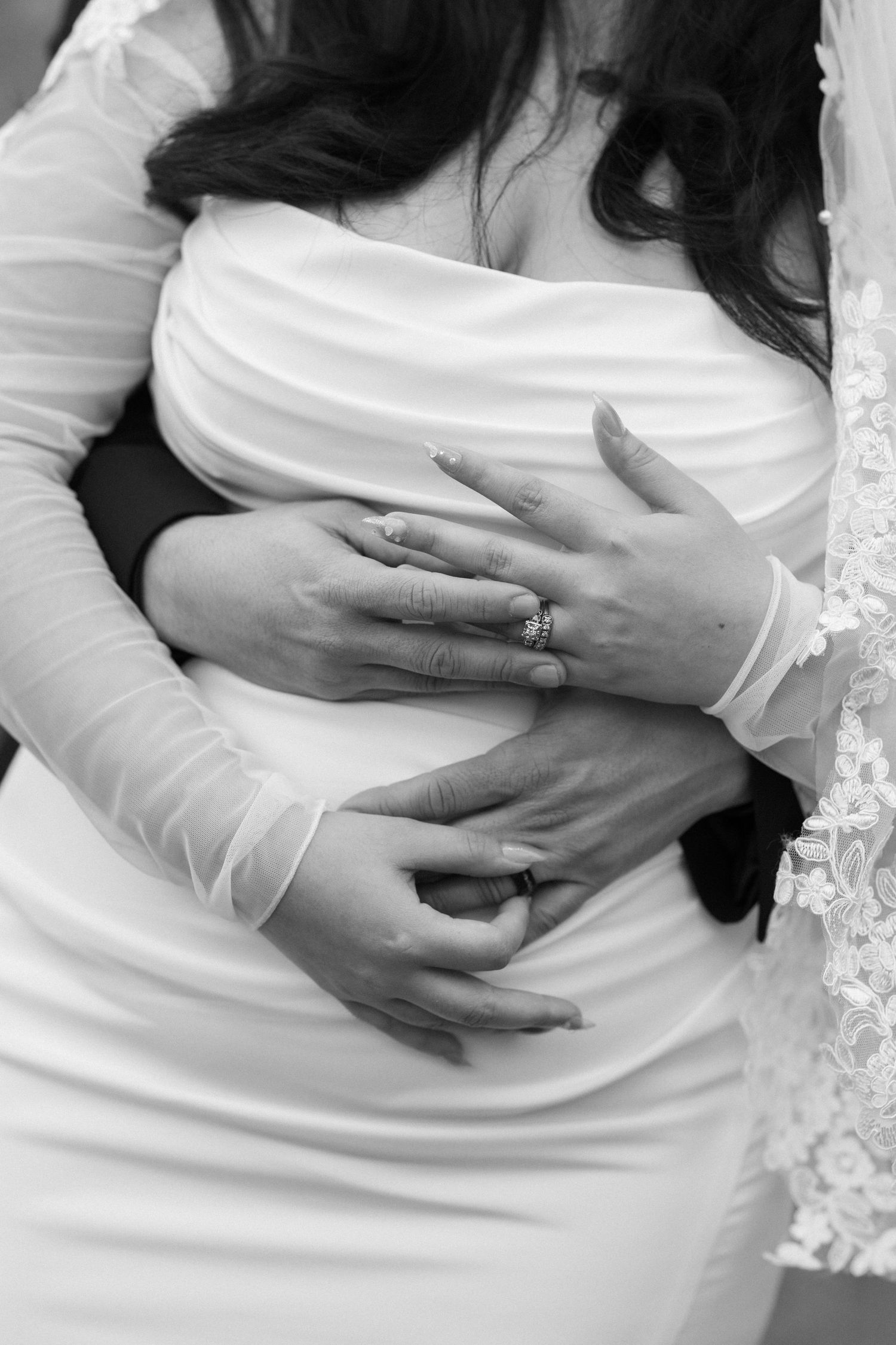 Close-up black and white photo of hands wearing rings embracing a white dress with lace details.