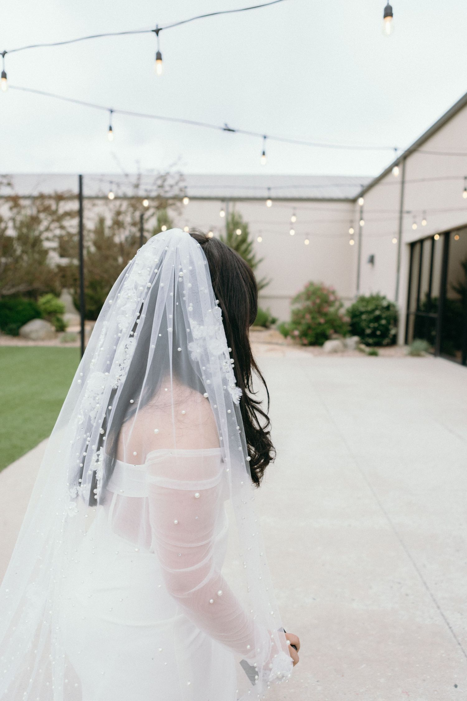 A bride wearing a delicate beaded veil with strings of lights overhead at an outdoor venue.