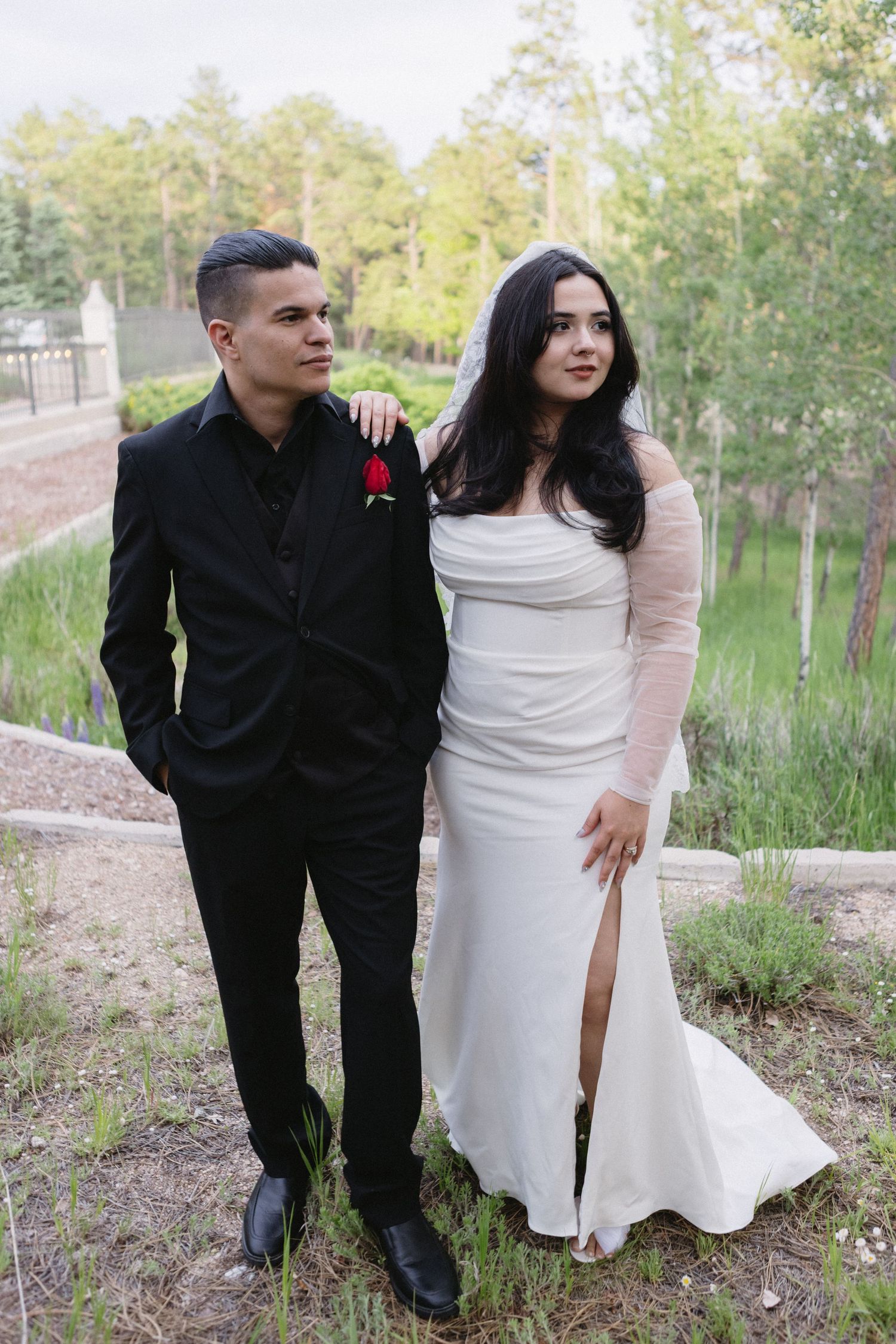 Two individuals in formal attire stand together outdoors against a background of trees and greenery.