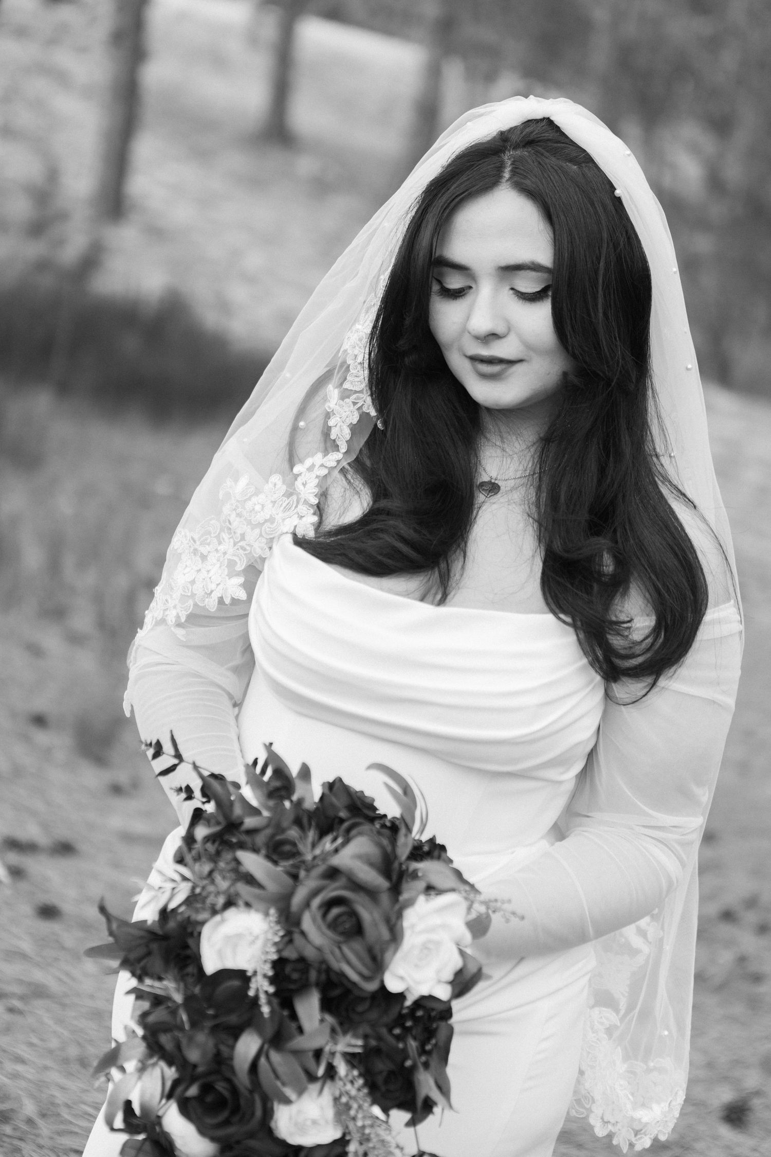 A bride in an elegant white dress with long sleeves and flowing veil holds a dark bouquet, captured in black and white.