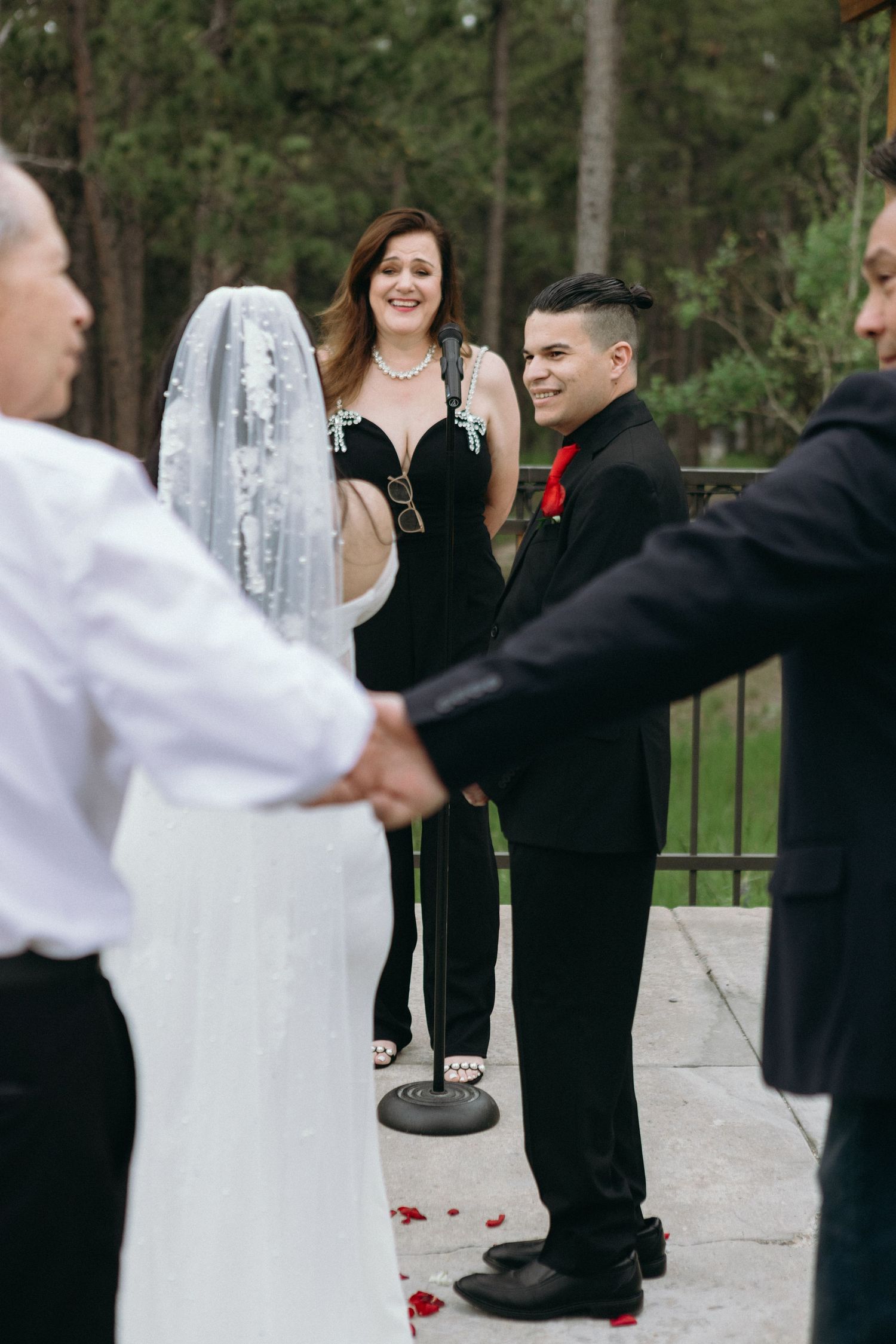 A wedding ceremony taking place outdoors with people in formal attire standing at an altar area with rose petals on the ground.