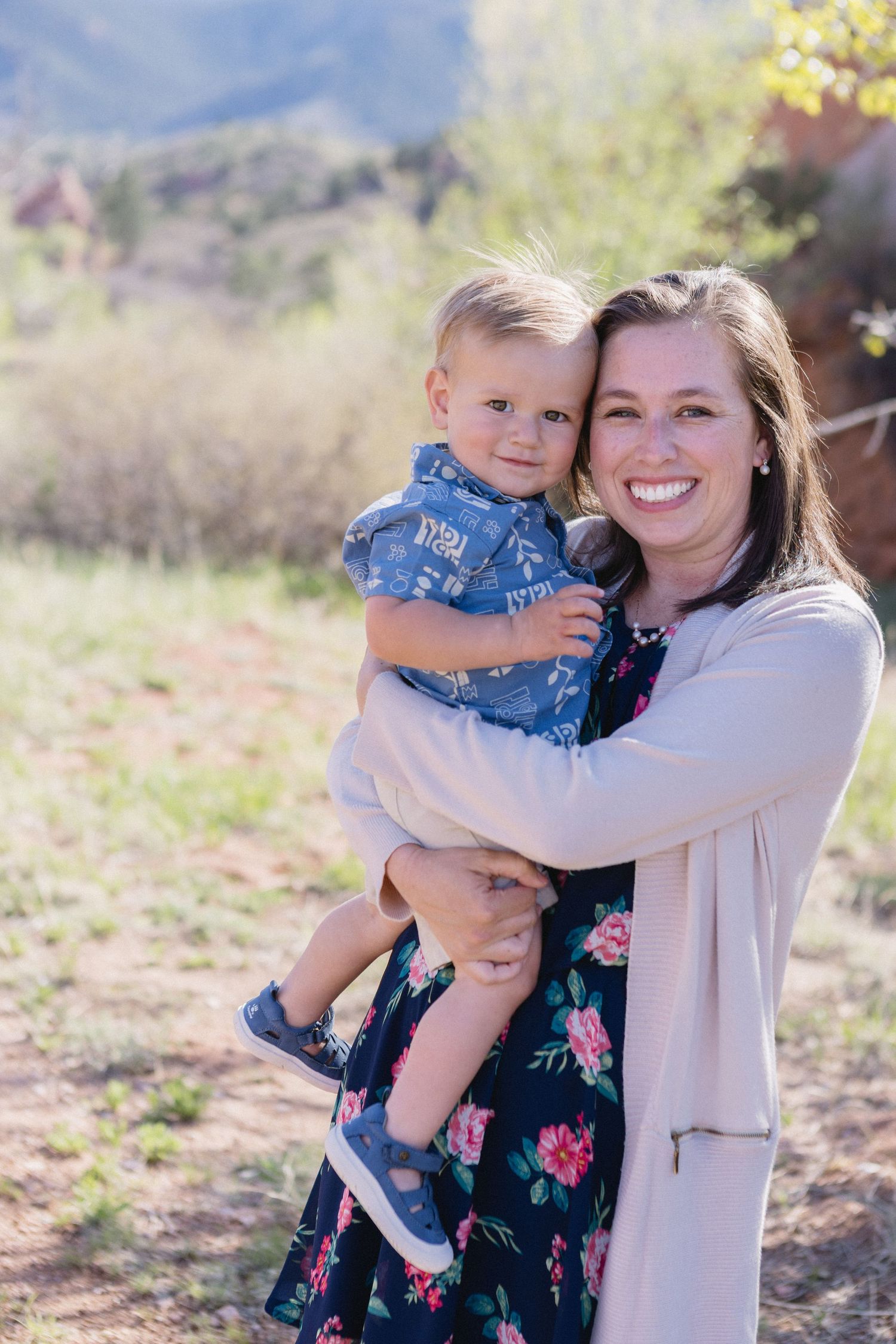A woman in a floral dress and cardigan holds a smiling toddler in denim outdoors on a sunny day.