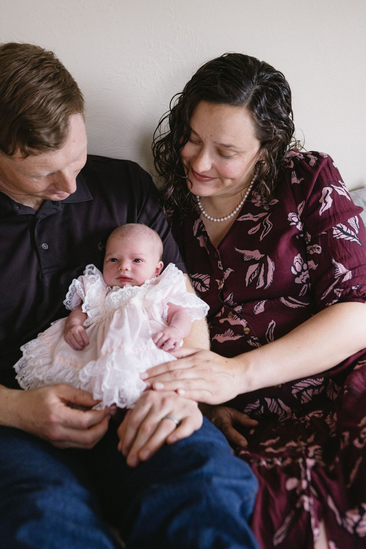 Family poses with newborn baby wearing white christening gown while sitting together indoors.