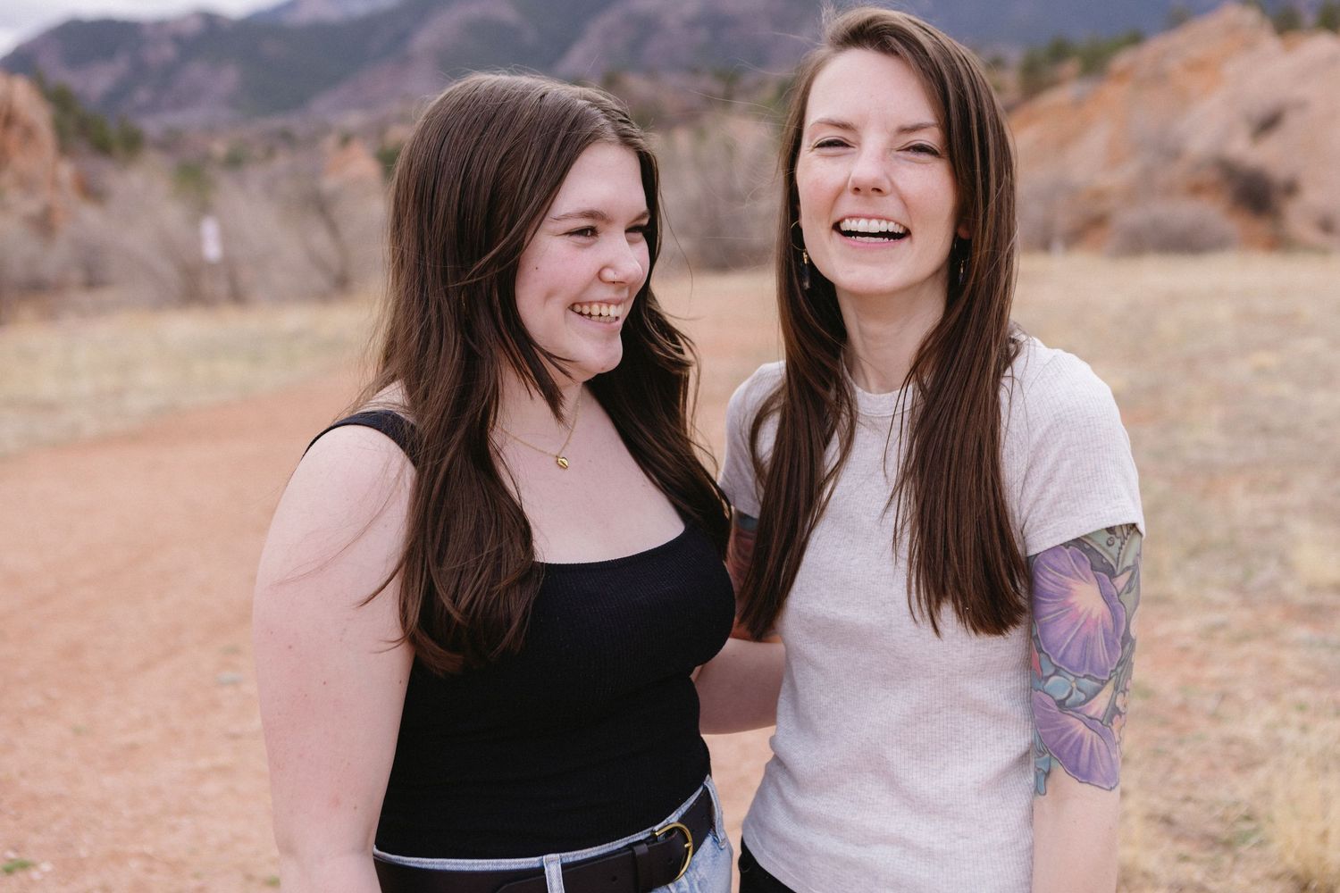 Two friends share a laugh together on a dirt path with rocky mountains in the background.