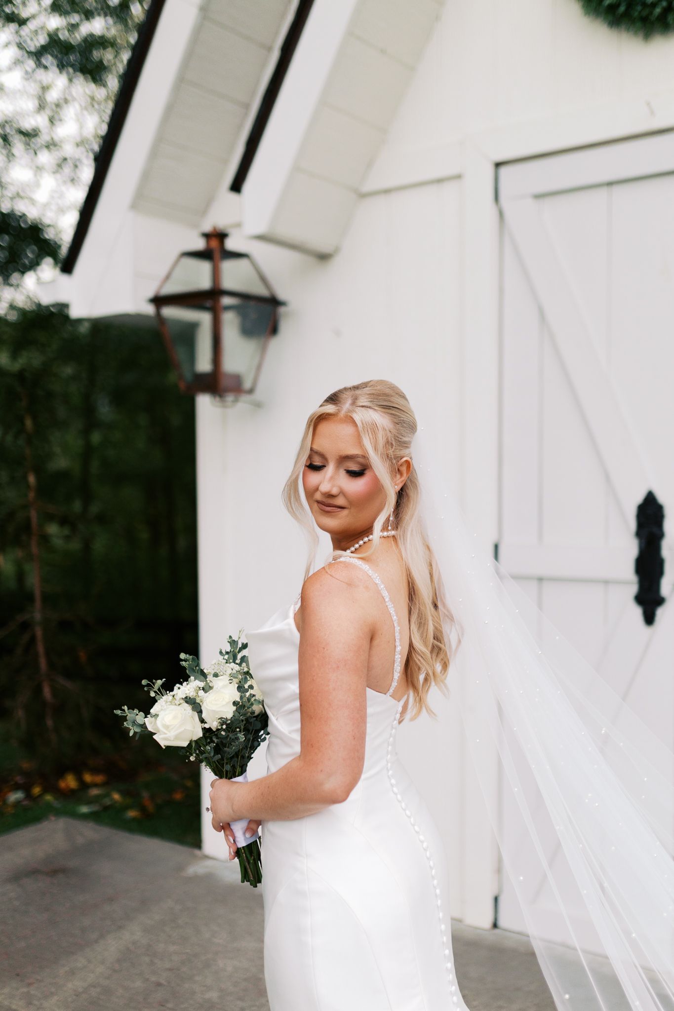 A bride in a white wedding dress poses elegantly against a white barn wall with rustic black hardware.