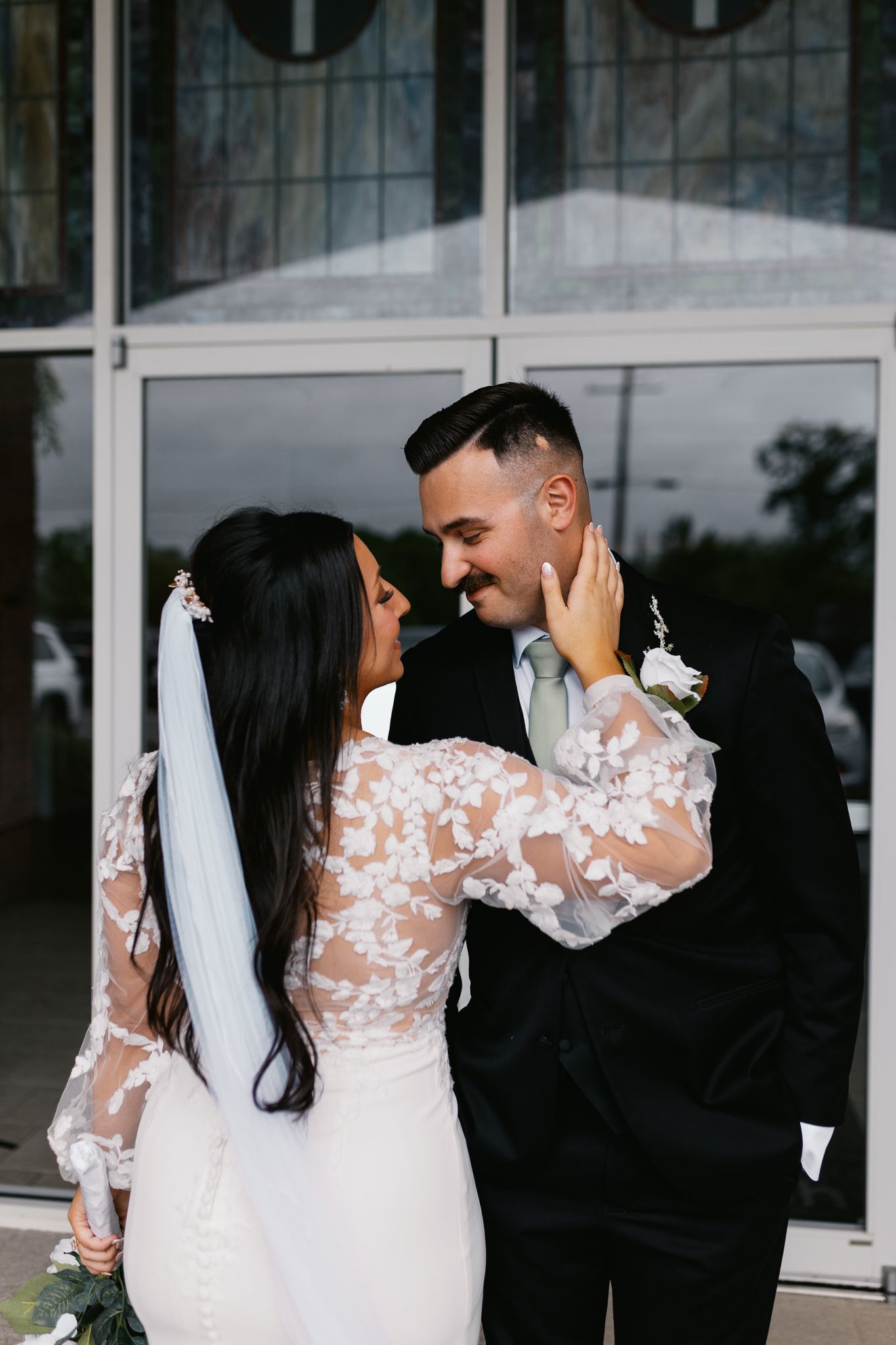 A couple shares an intimate moment in wedding attire against a glass building exterior.