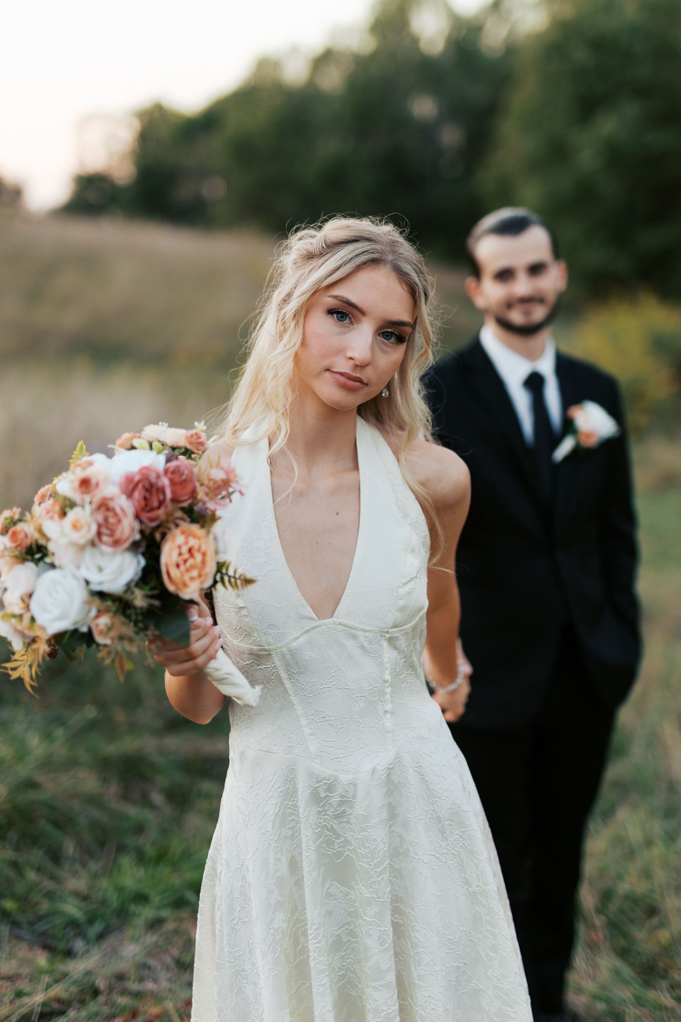 Bride holding bouquet in white dress with groom in background during outdoor wedding.