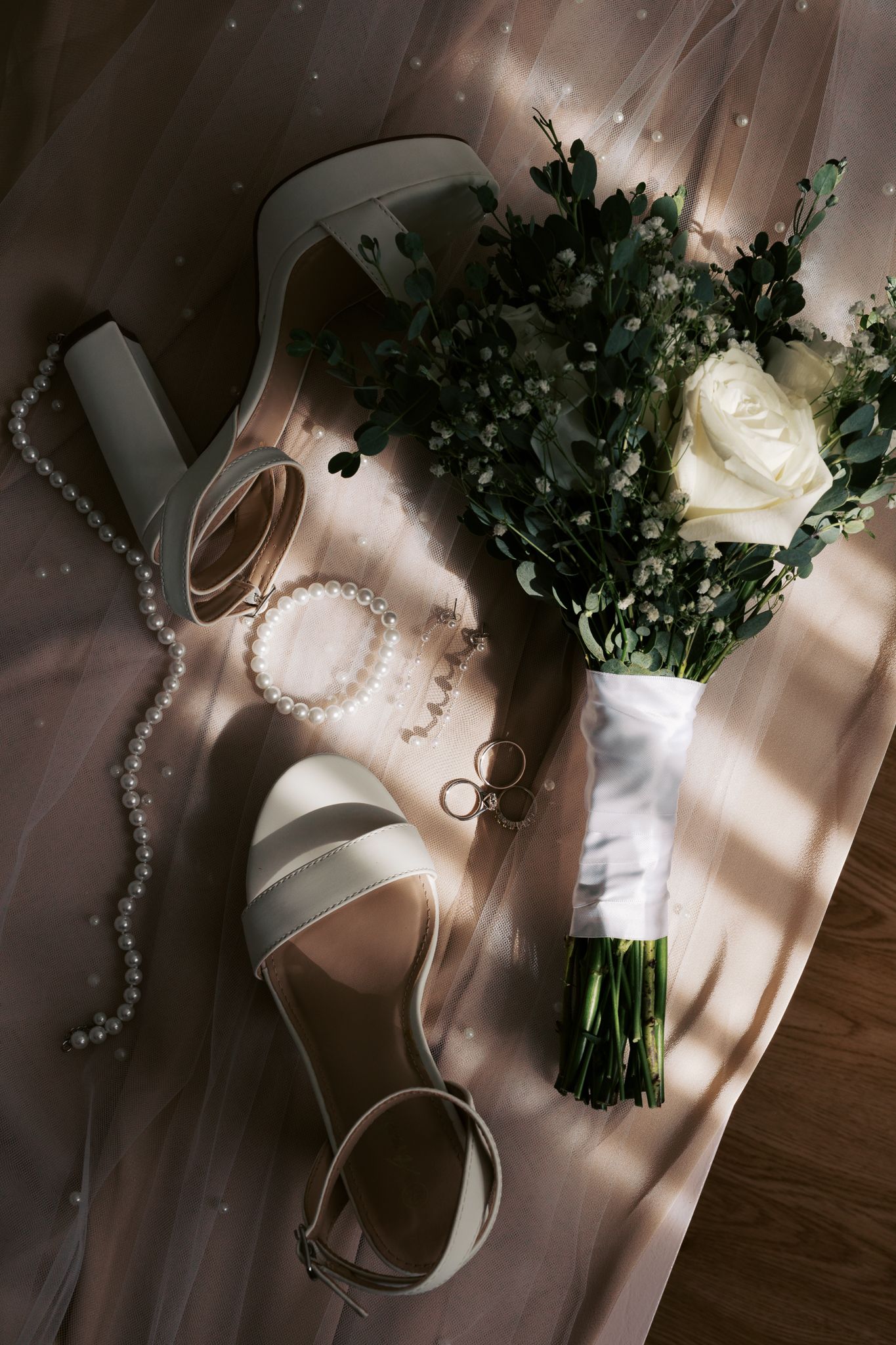 Wedding details including white roses, pearls, and ribbons arranged on a wooden surface in natural light.