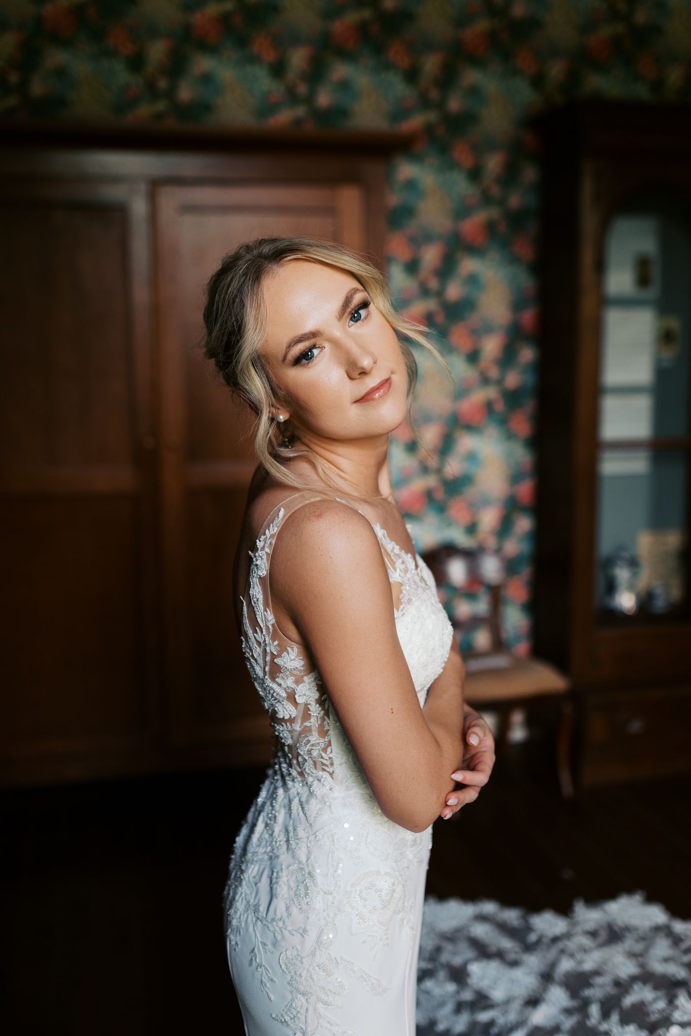 A bride in a lace wedding dress poses elegantly against a dark floral backdrop.