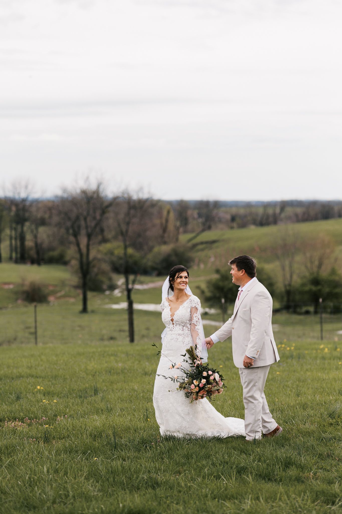A romantic wedding couple poses together on a grassy hillside in white attire during their outdoor photoshoot.