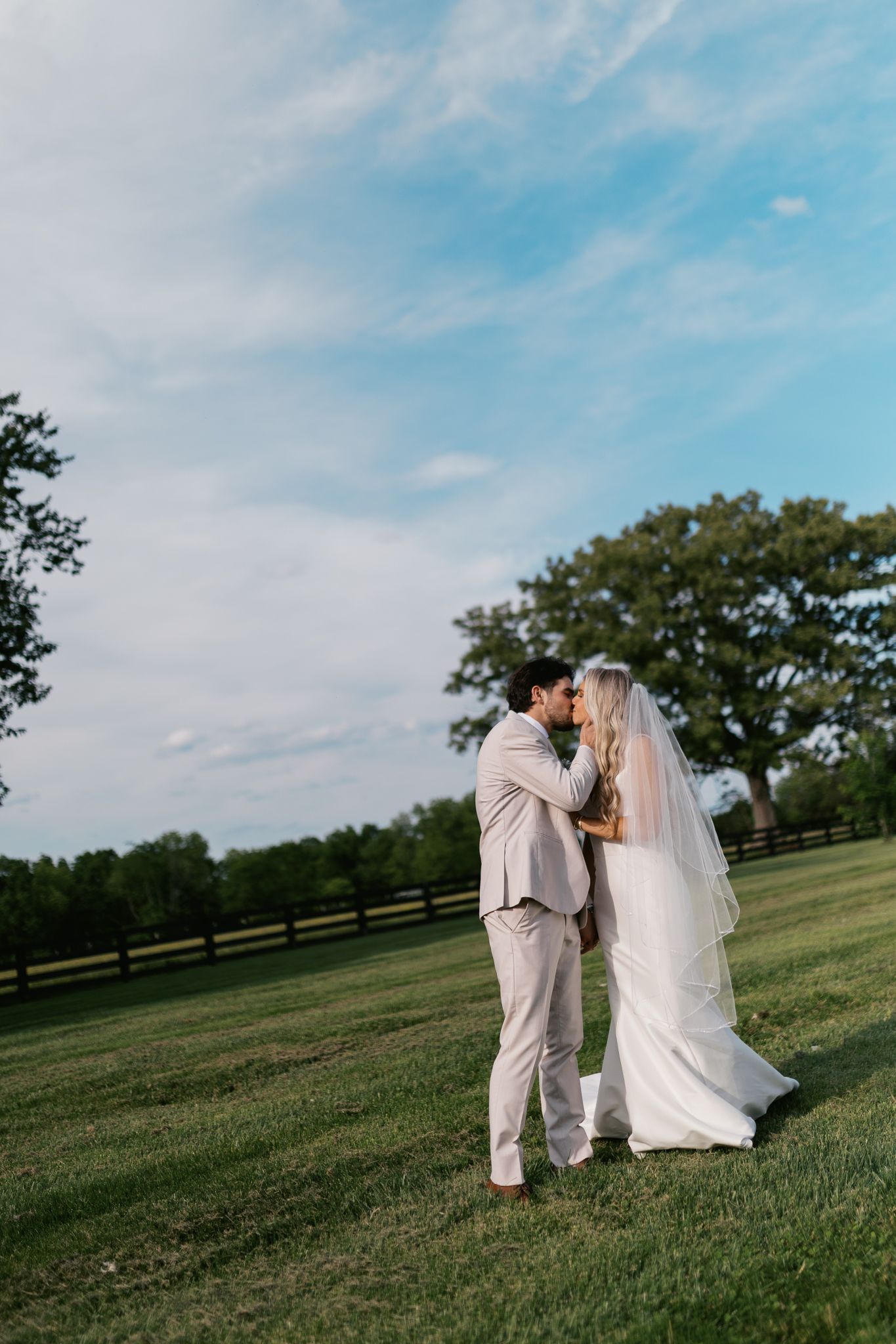 A couple shares an intimate moment in a scenic field with blue skies and wooden fencing.