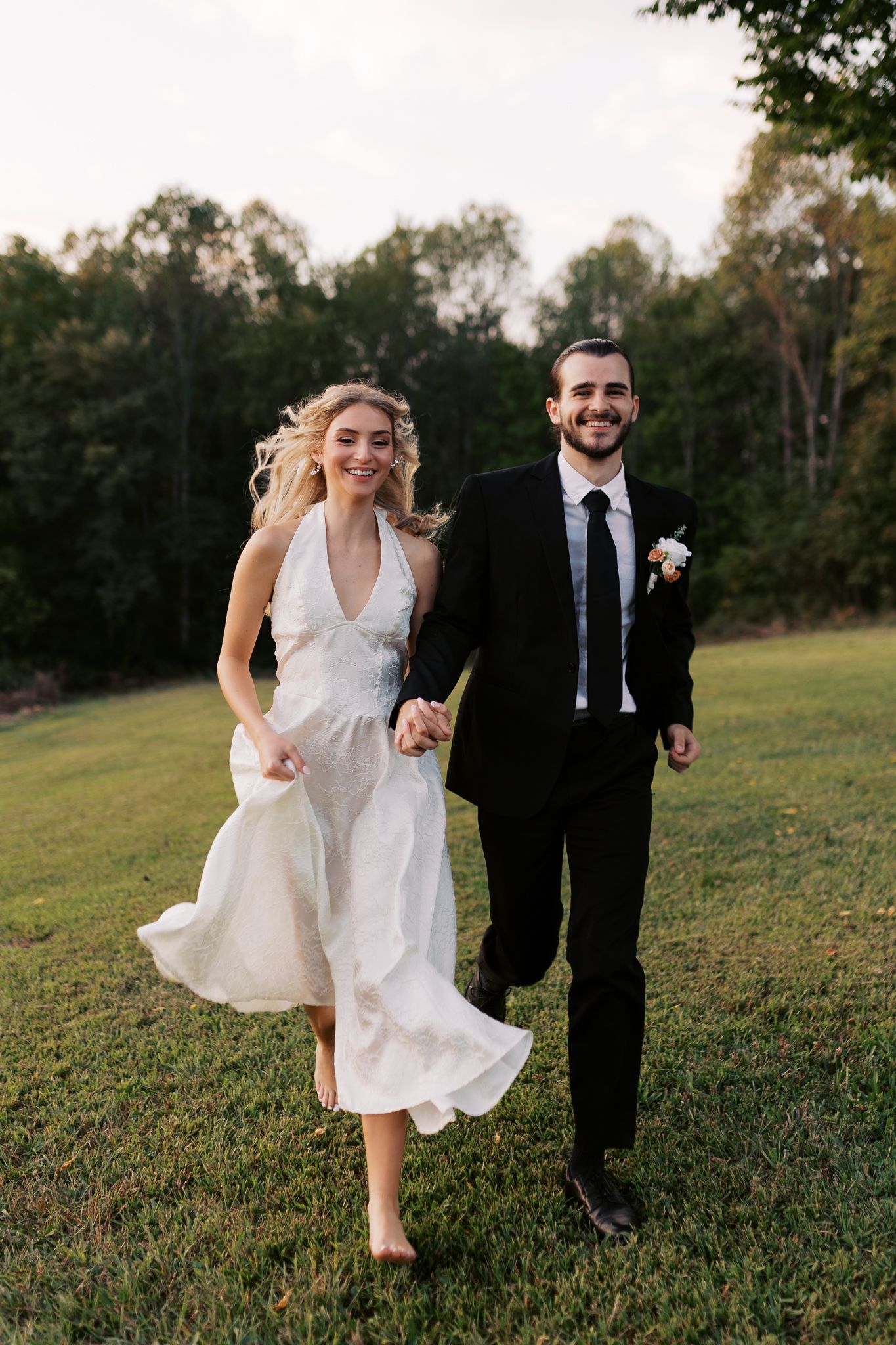 Couple running through a grassy field in wedding attire at sunset.