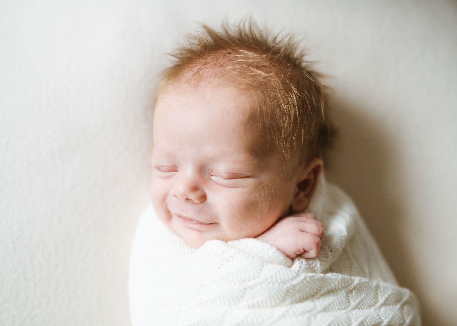 Peaceful newborn baby wrapped in soft white blanket smiles gently while sleeping against white background.