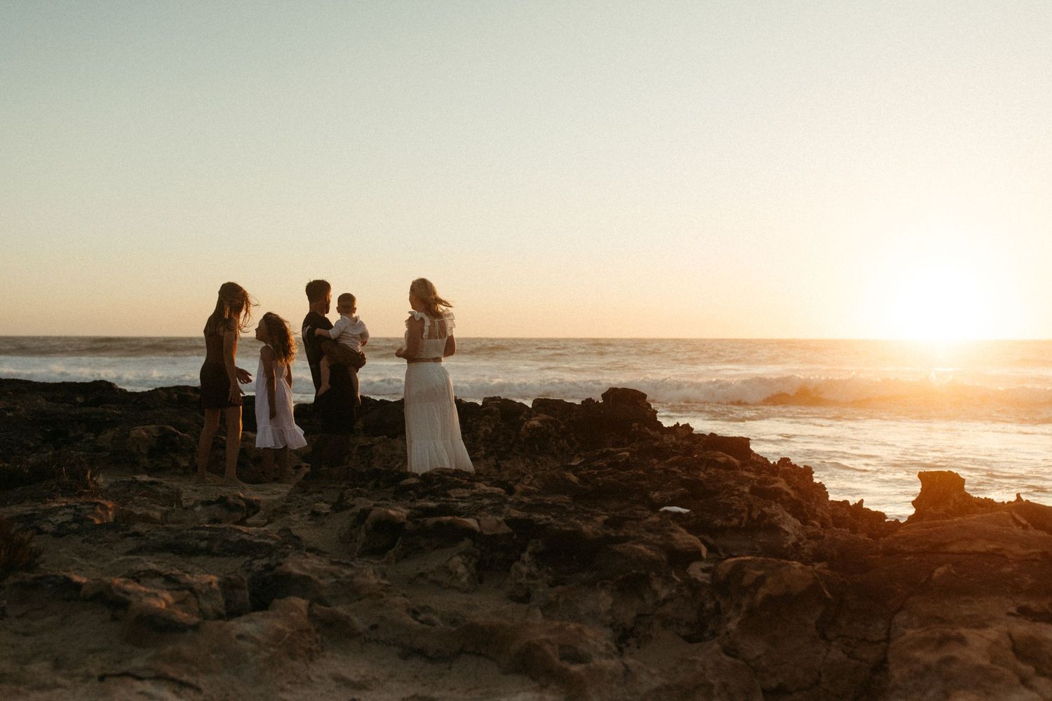Silhouettes of people standing on rocky coastline at sunset while ocean waves crash against shore.