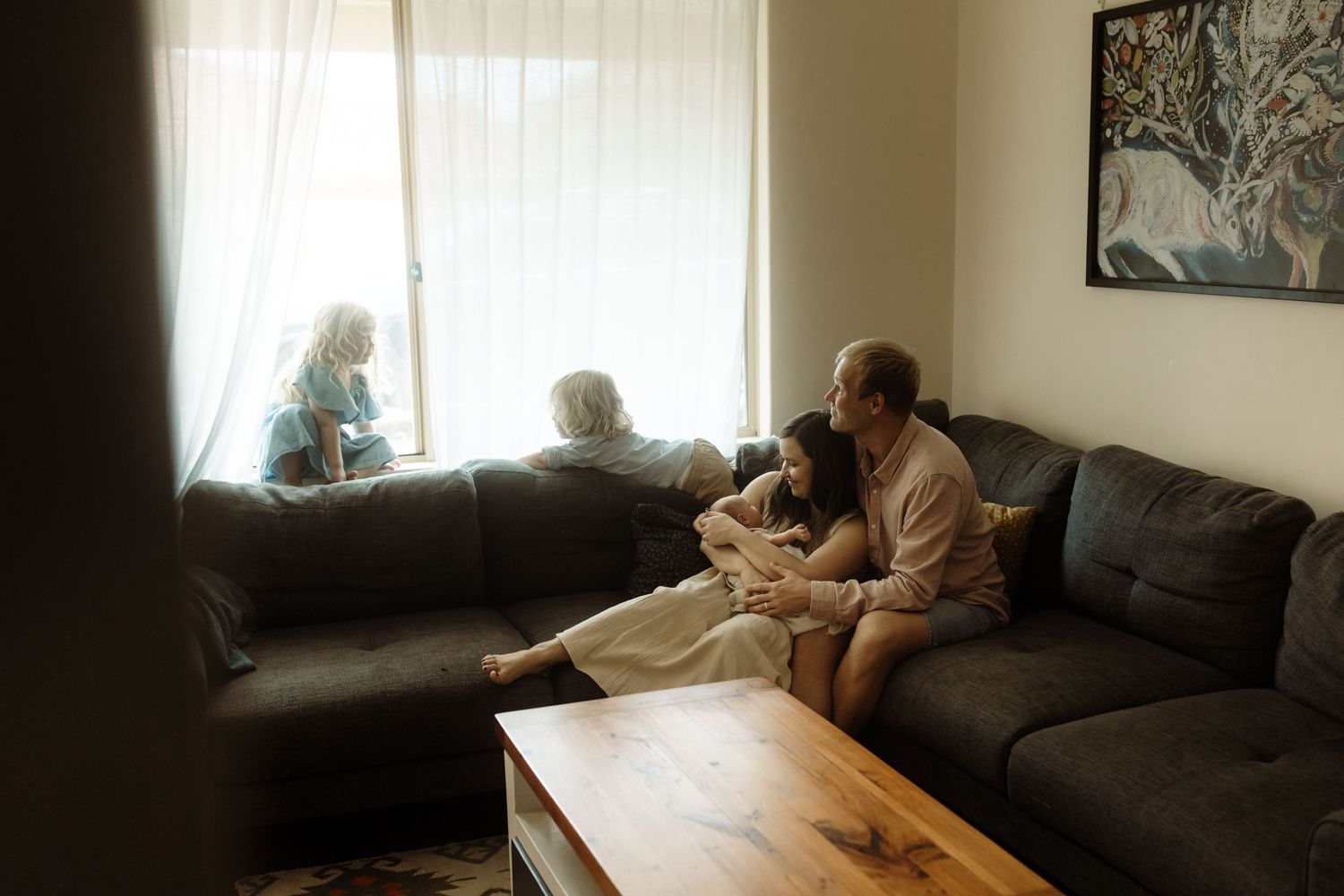 Two people relaxing on a grey sectional couch in a cosy living room with natural light streaming through windows. In home newborn session.