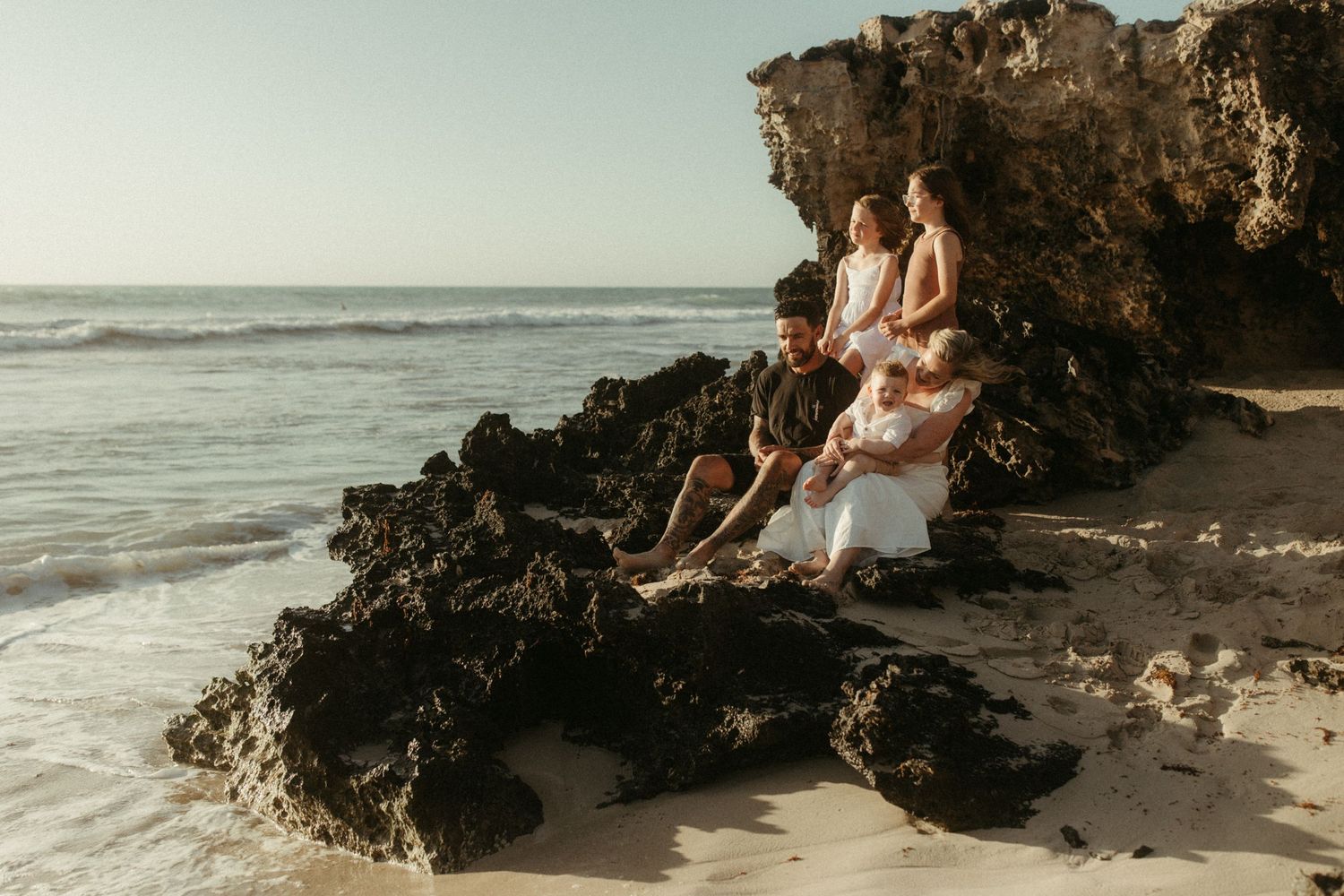 Two people sit together on volcanic rocks at the beach during sunset with waves crashing in the background.