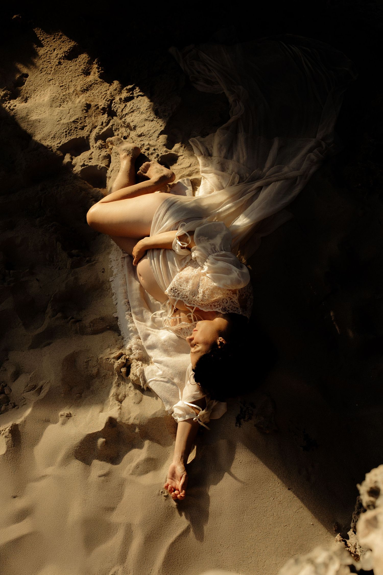 Ethereal figure in flowing white dress lying on sand near rock formations in dramatic lighting.