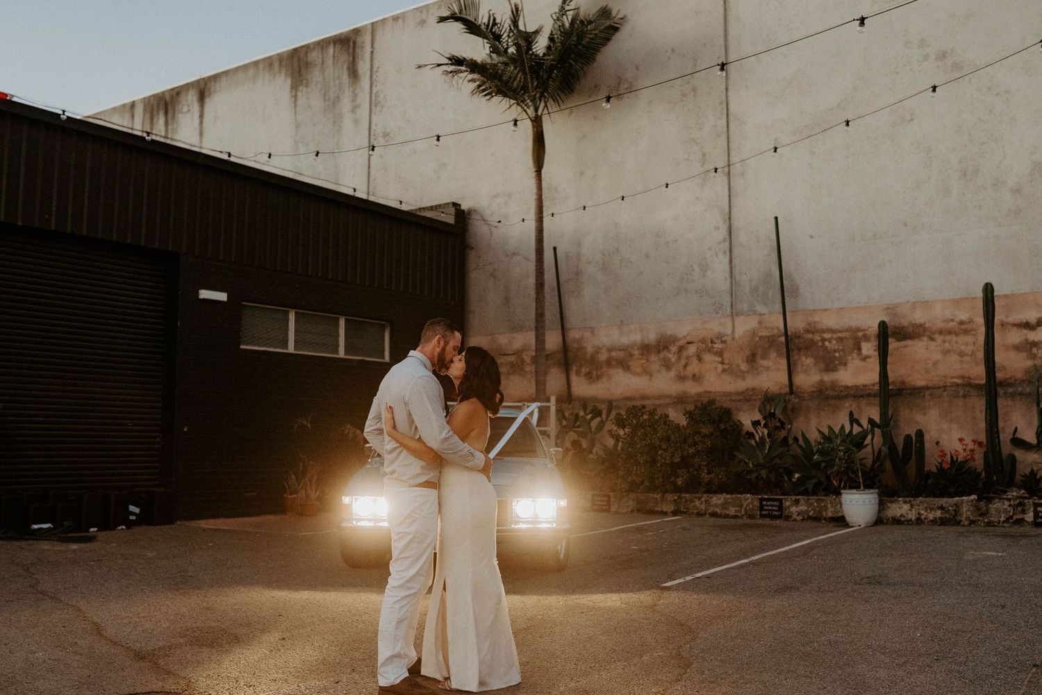 Couple dancing in white outfits at sunset with car headlights illuminating them.