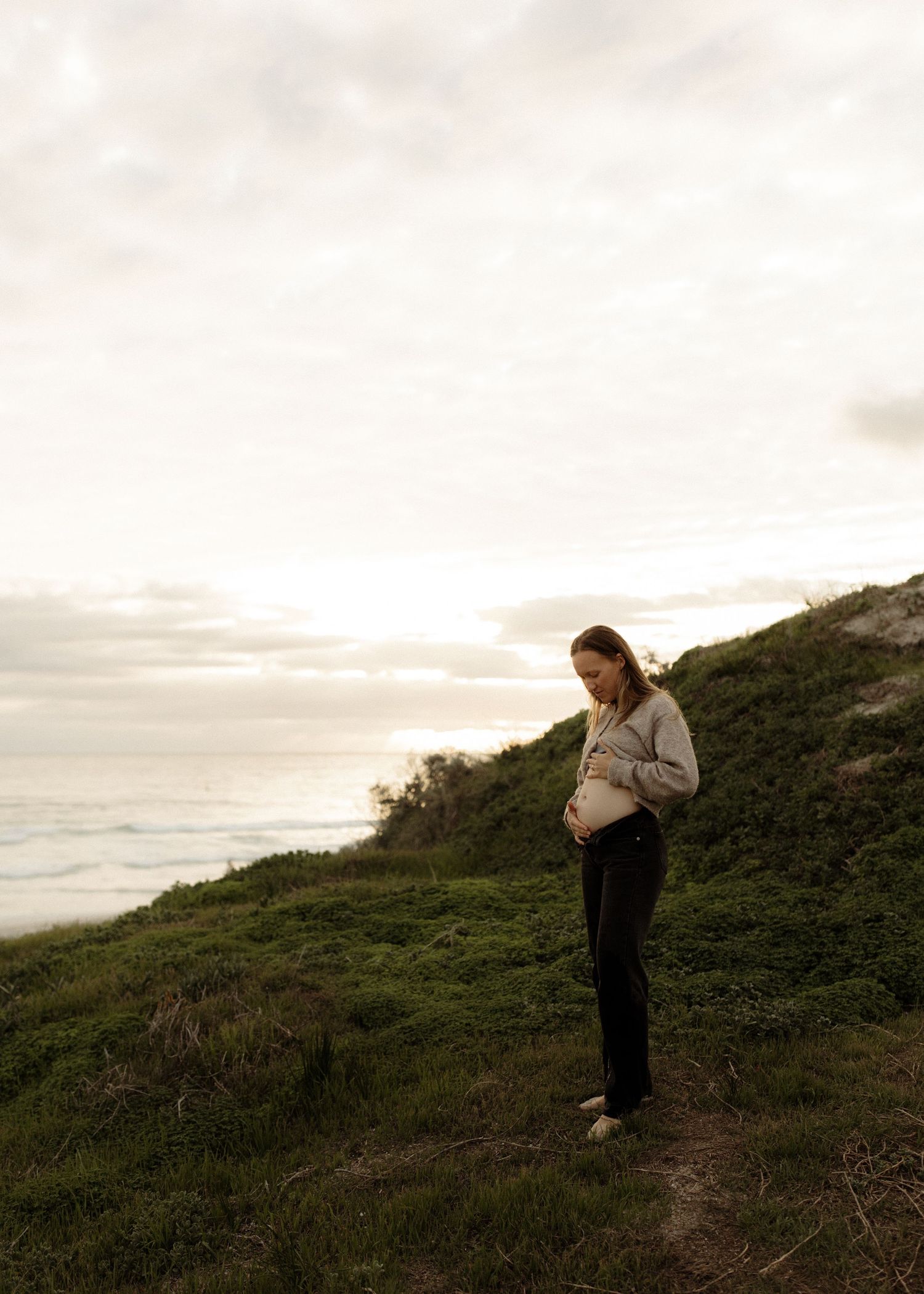 A person in dark clothing walks along a steep coastal path at dusk with an ocean view in the background.