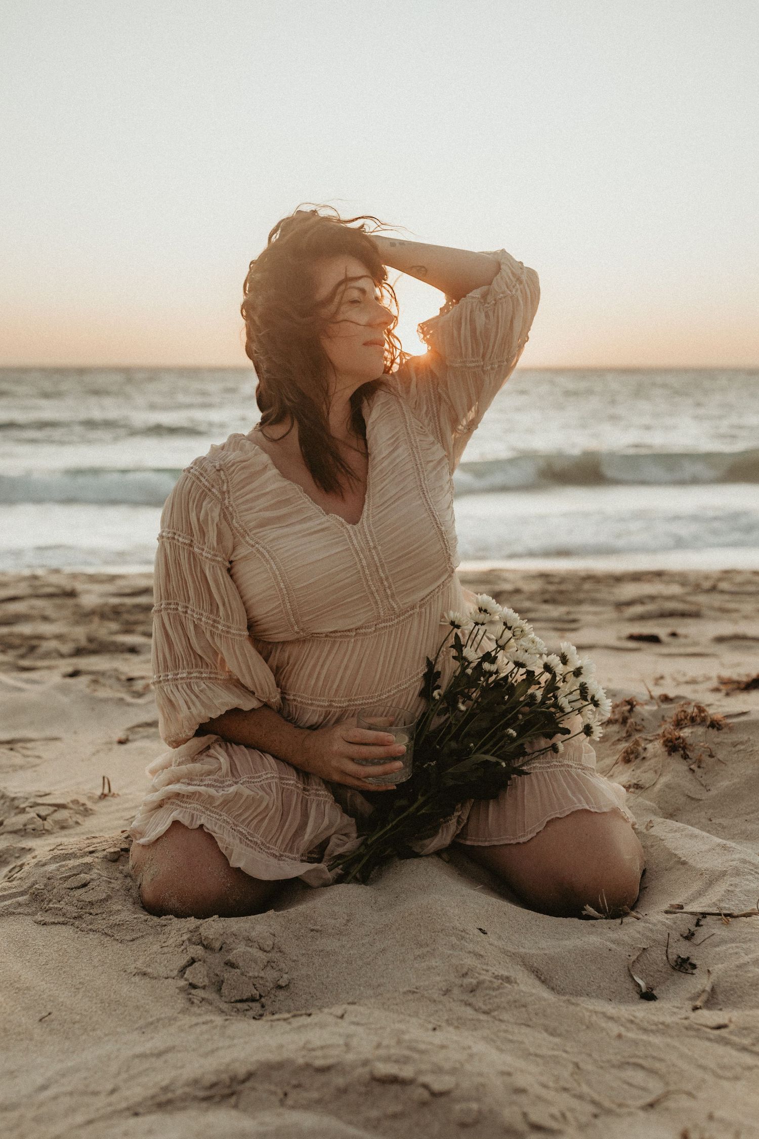 Woman in white dress poses with flowers on beach at golden hour sunset. Courtney Healy Photography Yanchep Perth