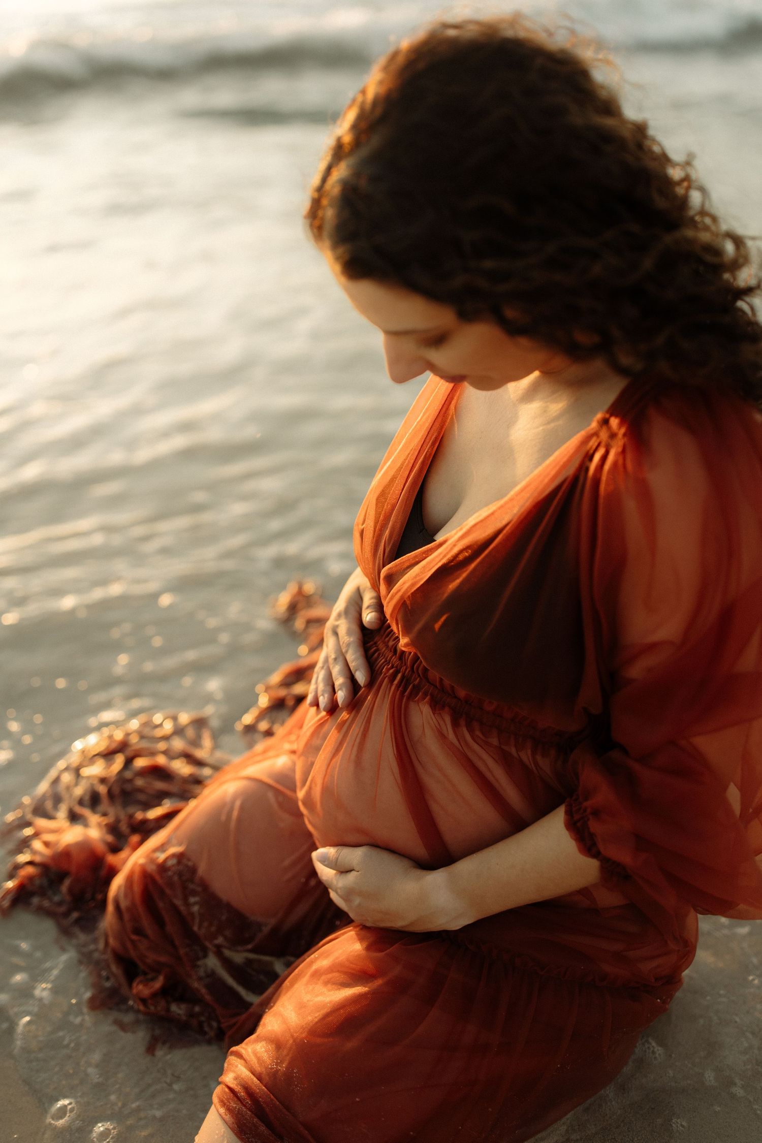 Maternity photo of woman in flowing red ochre dress sitting by the ocean at golden hour in Perth, Western Australia.