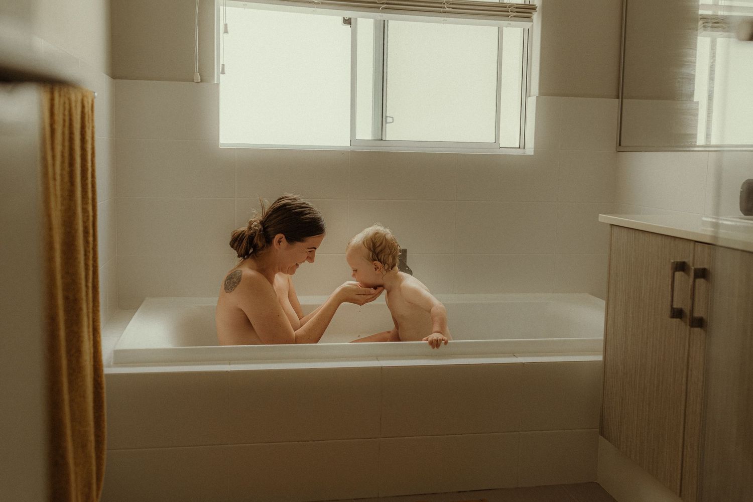 Tender bathtub moment between parent and child in minimalist bathroom setting. Motherhood photography
