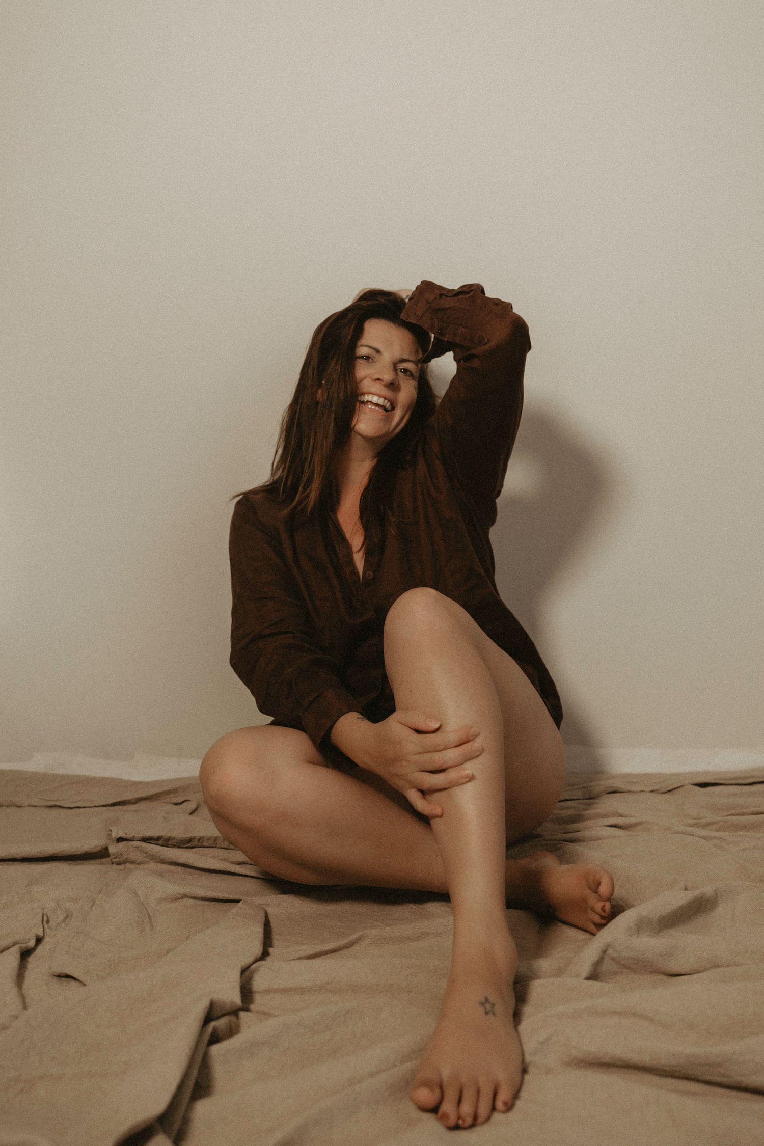 Woman in brown shirt laughing while sitting on beige blanket during indoor photoshoot.