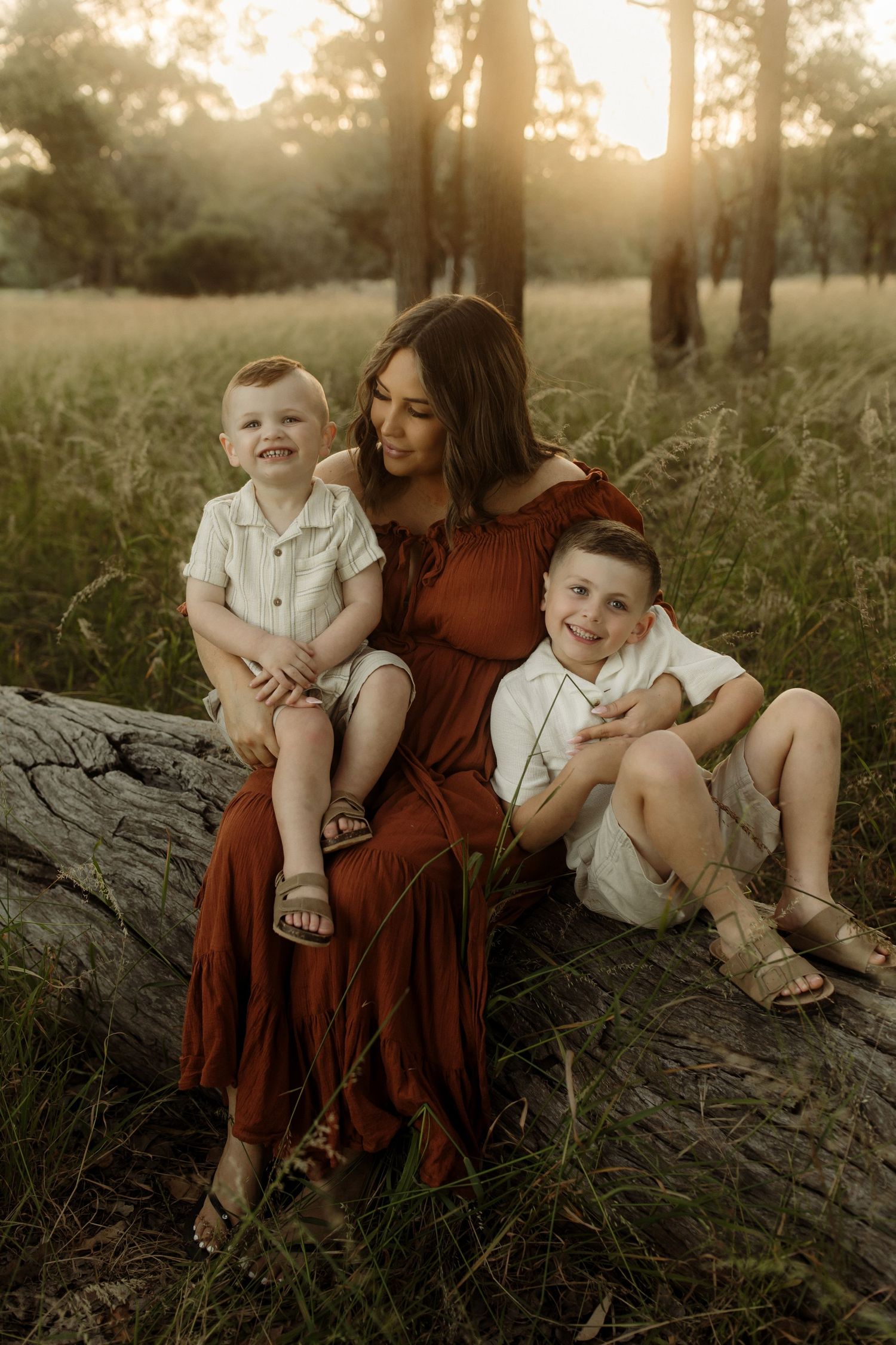 Warm sunset backlit family portrait on fallen log in grassy field with tall trees in background.