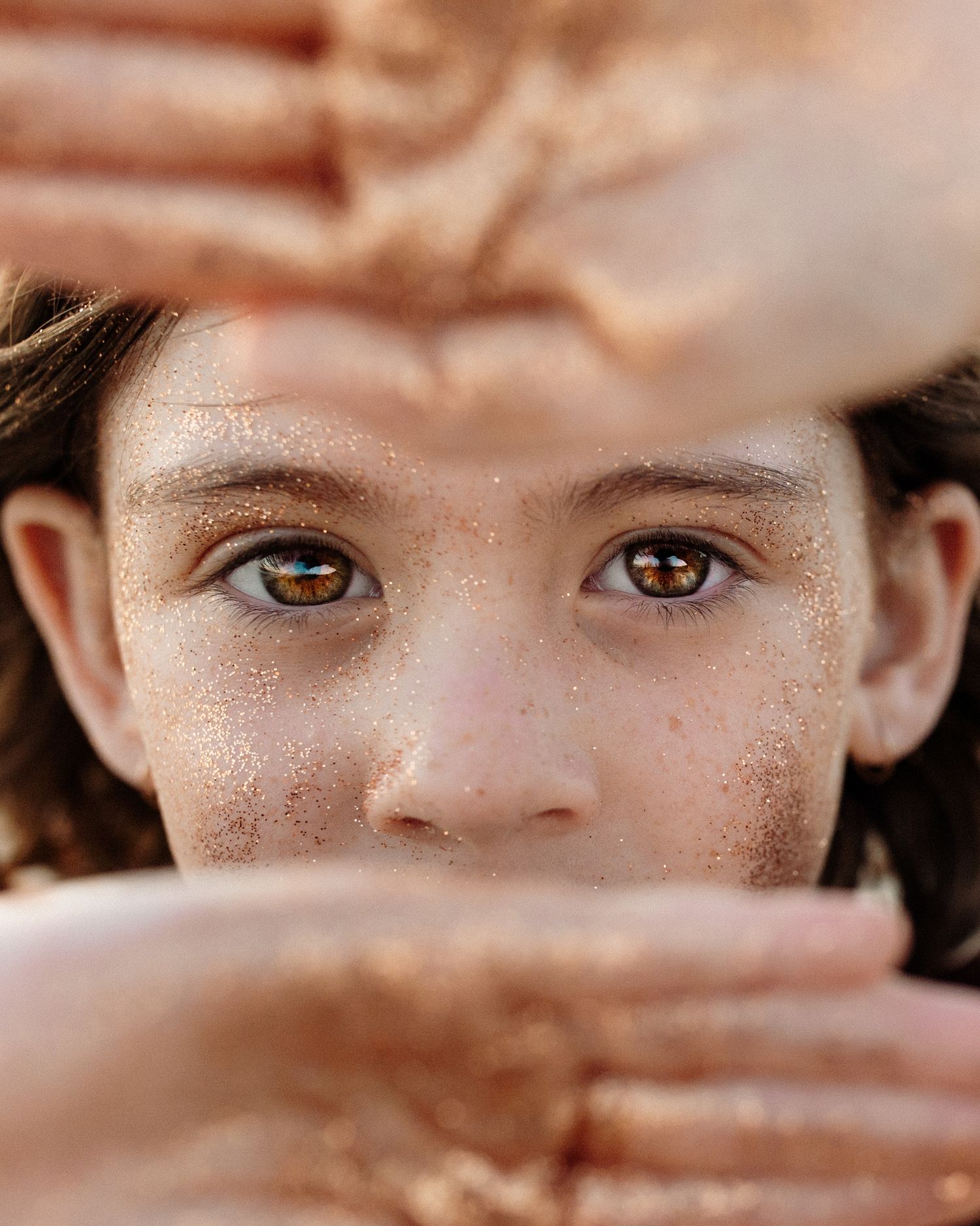Artistic shot showing intense hazel eyes framed by hands with a soft blurred background.