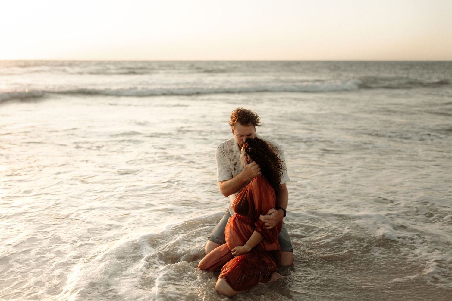 Couple sharing tender moment while standing in gentle ocean waves at sunset. Maternity Session