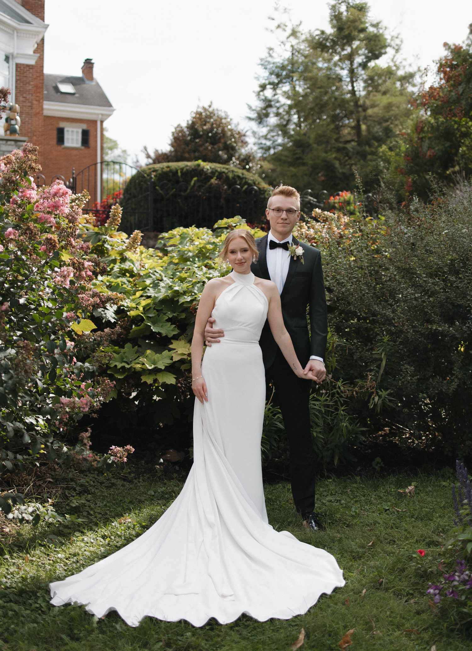 A couple poses in a garden setting with white wedding attire against lush greenery and flowers.