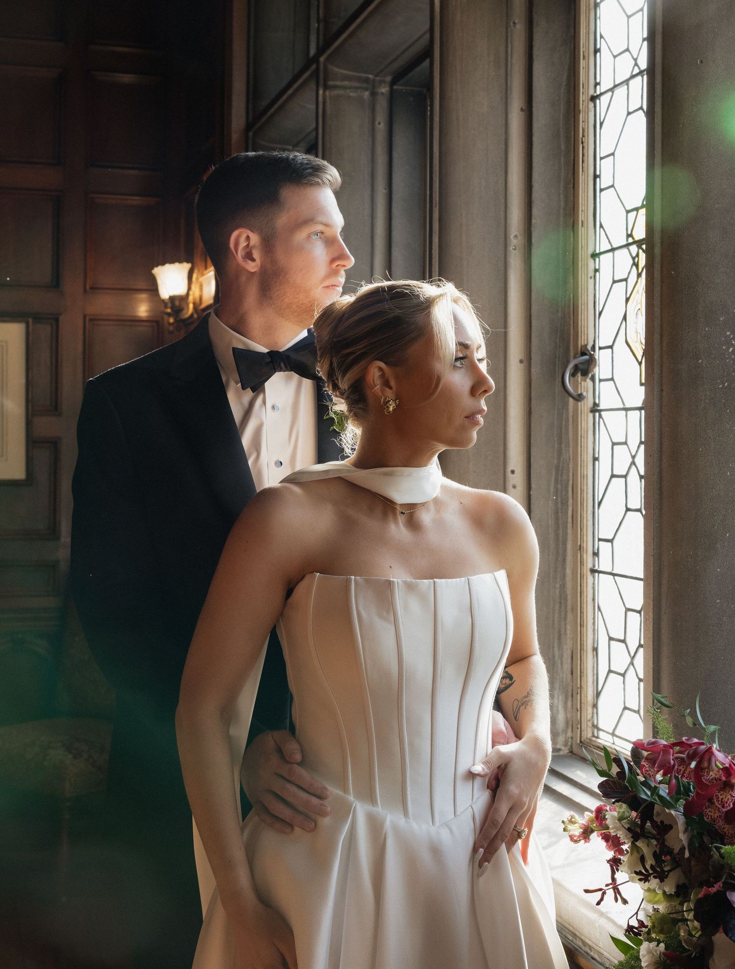 A couple in formal wedding attire stands by a window in a historic building with ornate details and natural light.