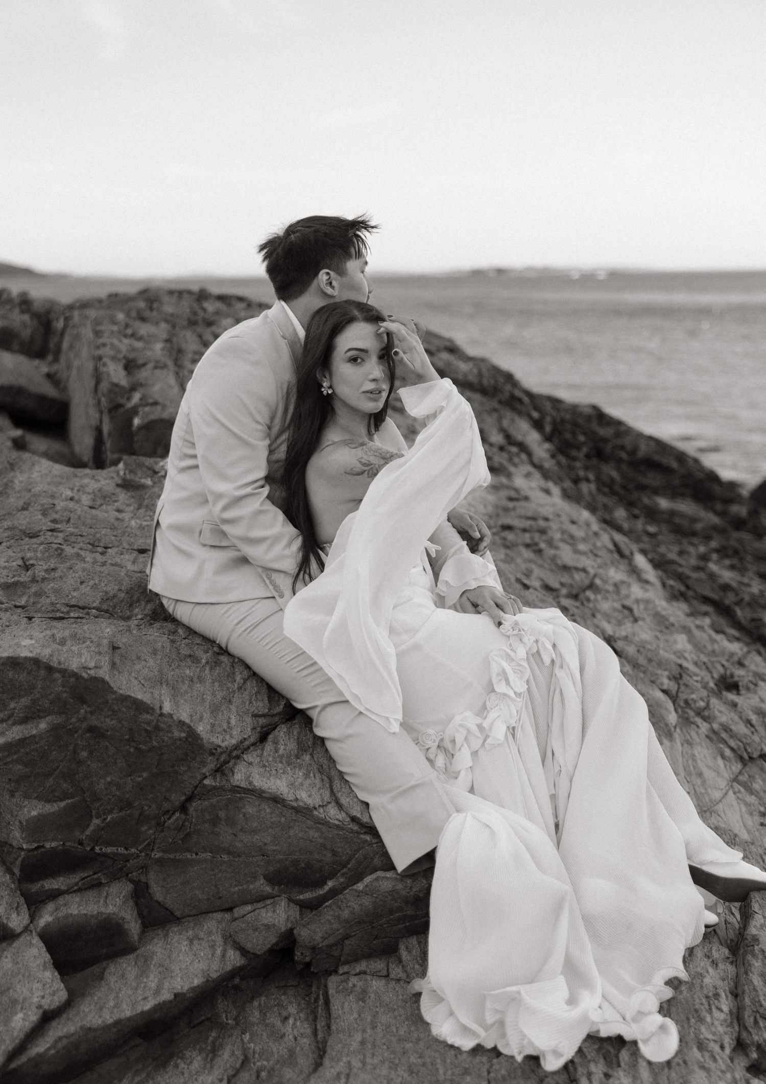 Black and white photo of a couple in wedding attire sitting on coastal rocks by the ocean.