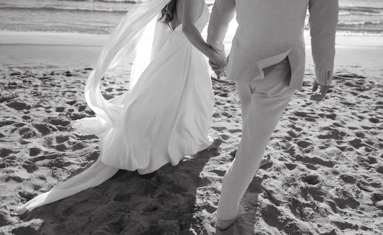 Wedding couple walking along sandy beach with flowing white dress.