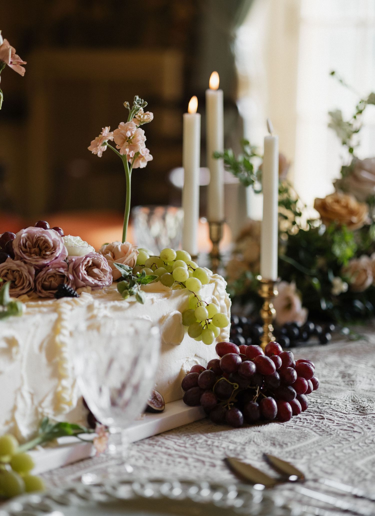 Elegant wedding cake with white frosting, fresh grapes, pink roses, and tall candles on a lace tablecloth.