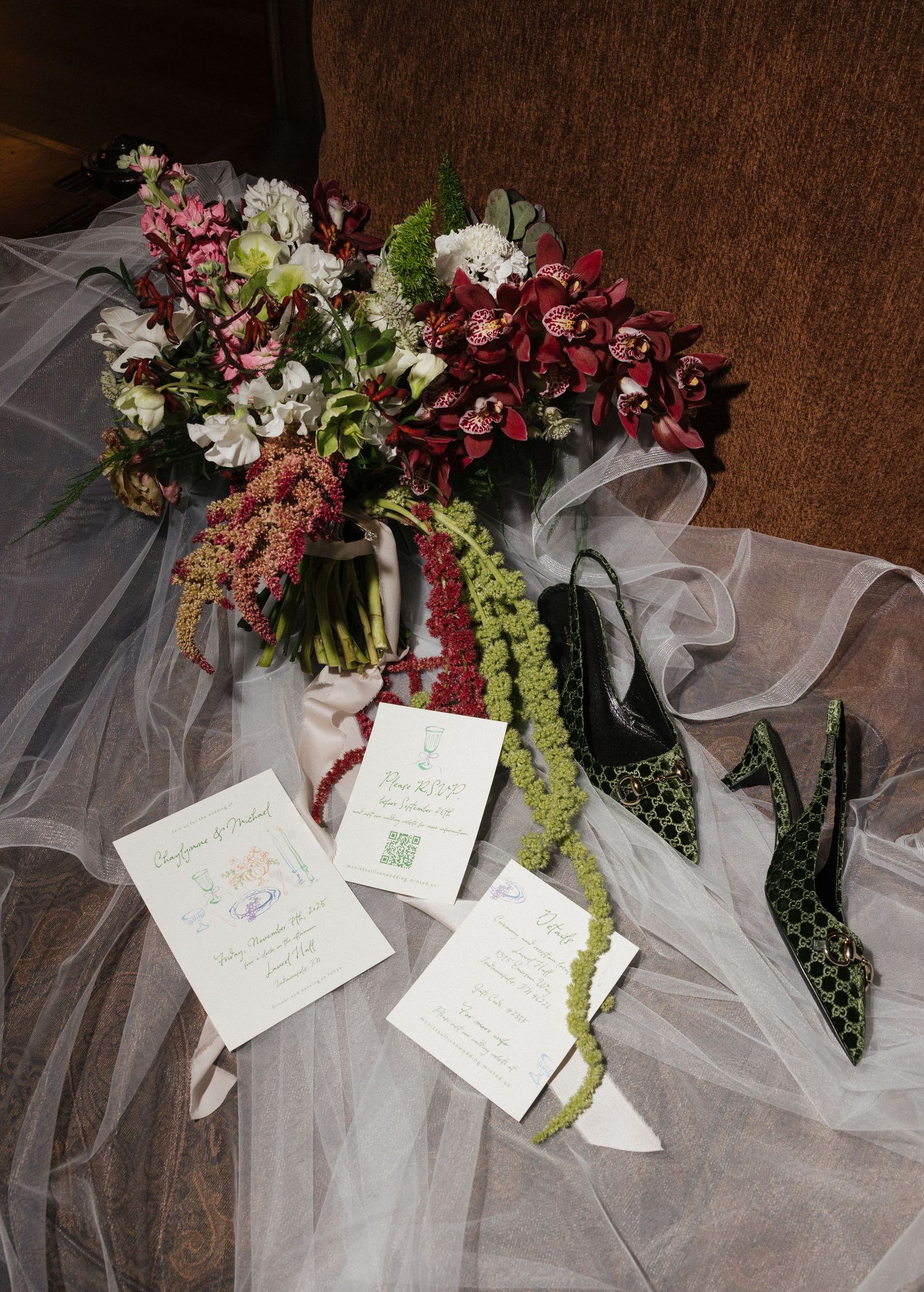 Bridal flat lay featuring burgundy orchid bouquet, green embellished heels, sheer veil, and elegant white wedding invitations.