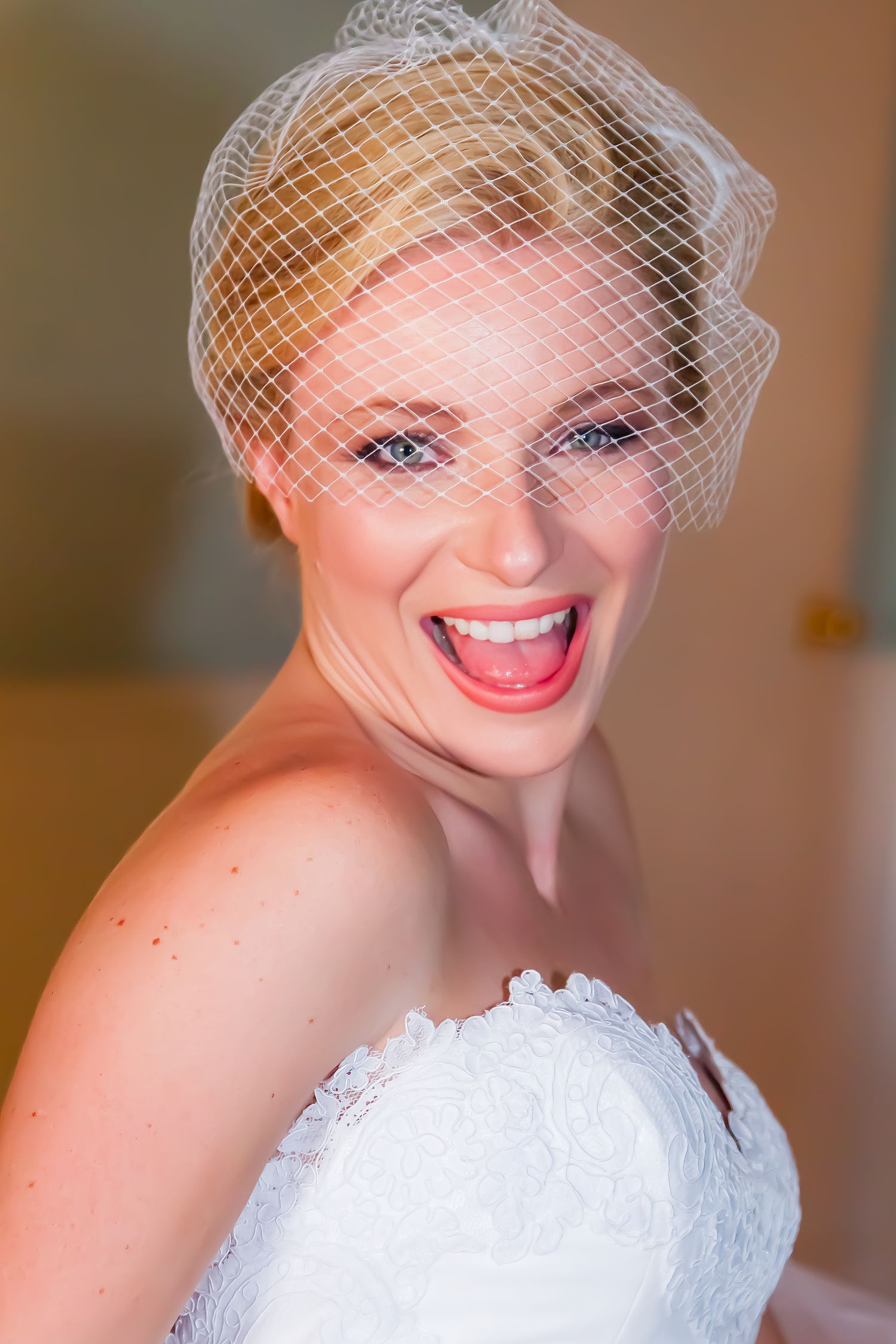 Radiant bride wearing a white strapless wedding dress and birdcage veil smiles joyfully in an indoor portrait.
