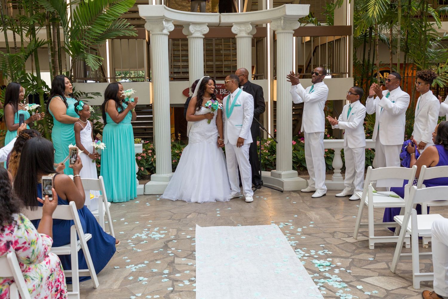 Elegant wedding ceremony with bride and groom at the altar, bridesmaids in teal dresses, guests seated in white chairs indoors.