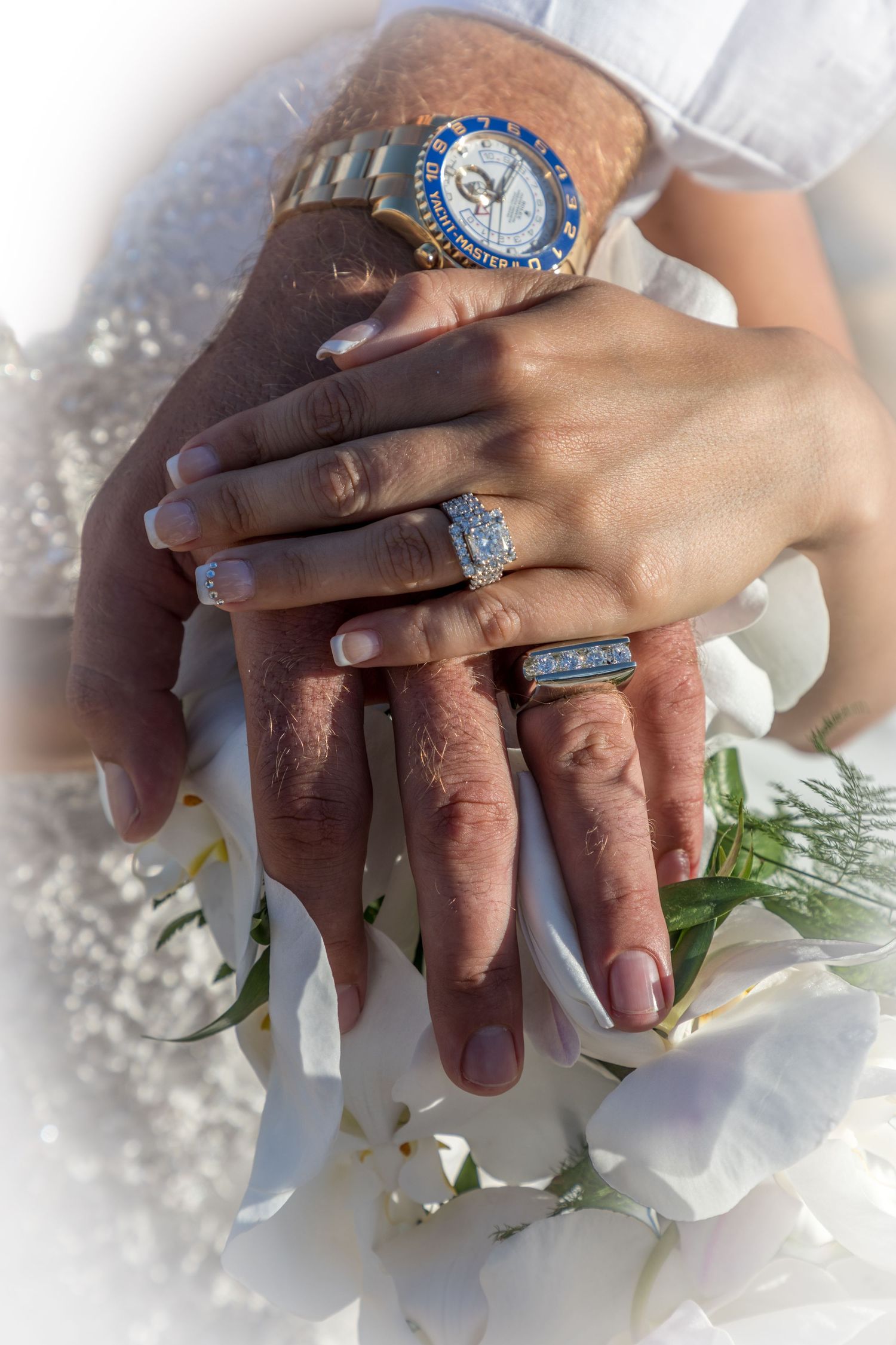 Newlywed couple displaying wedding rings and a luxury watch atop a white floral bouquet.
